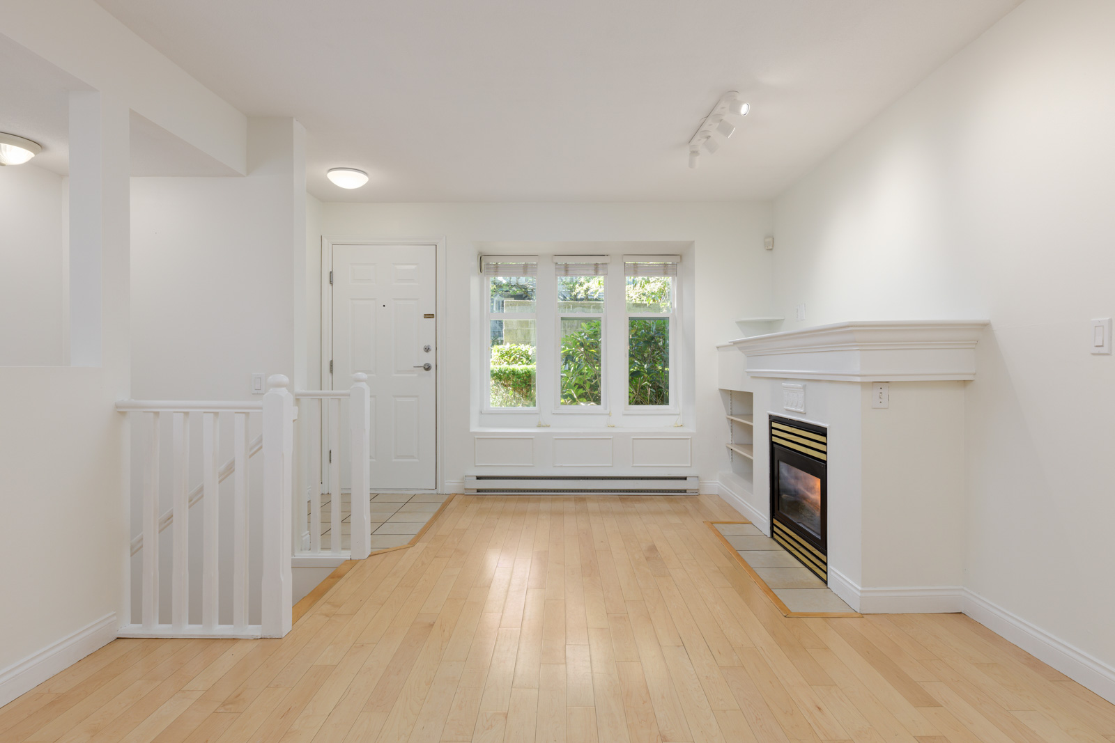 Bright, empty living room with light wood floors, white walls, a white fireplace on the right, three windows, and a staircase on the left near the front door.