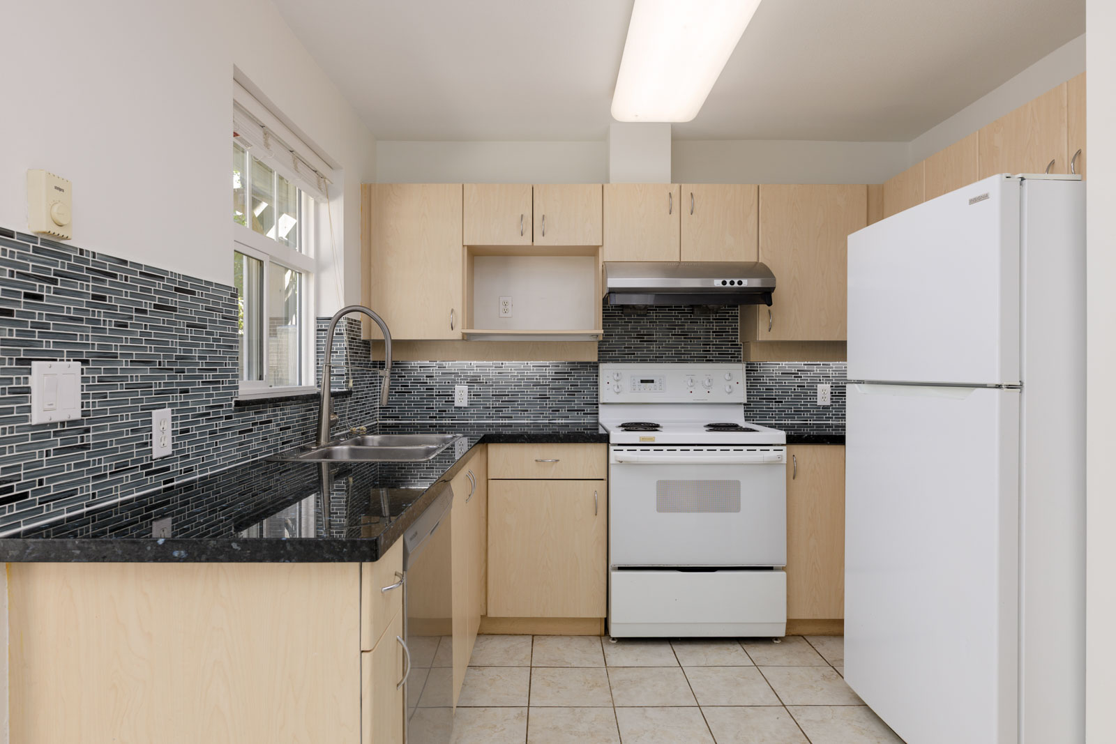 Modern kitchen with light wood cabinets, white appliances, black granite countertops, and a gray tile backsplash.