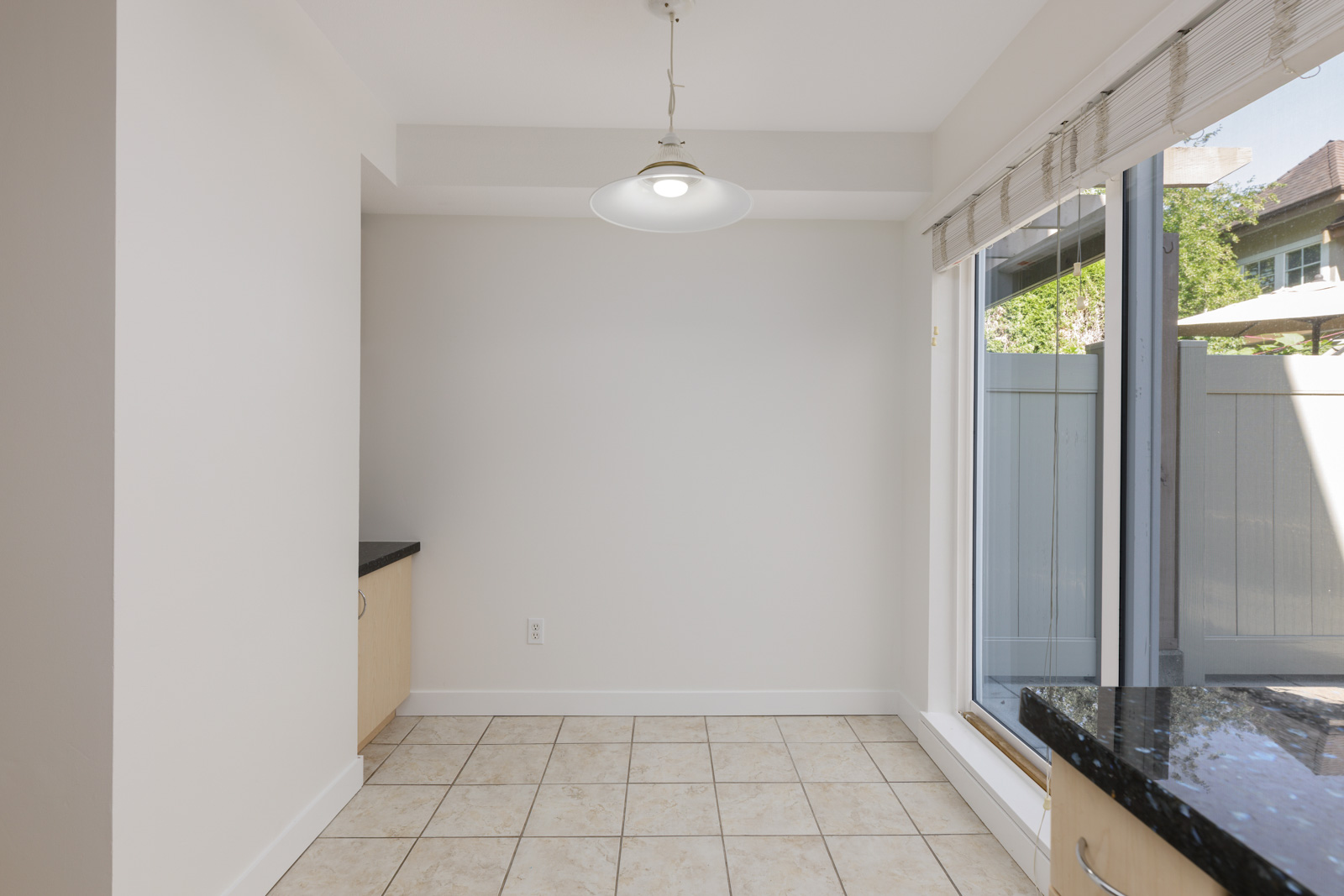 Bright, empty dining area with tiled floor, hanging light fixture, large window with blinds, and partial kitchen counter visible.