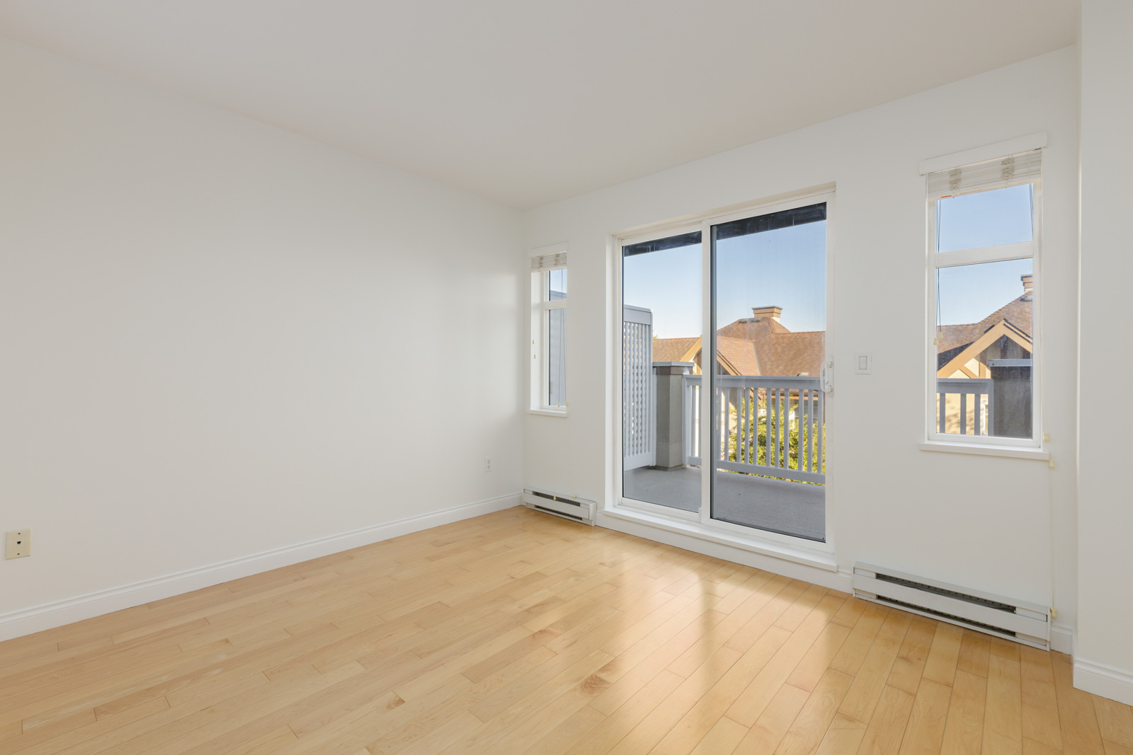 Empty room with light wood flooring, white walls, two small windows, and a glass door leading to a balcony with a view of neighboring rooftops.