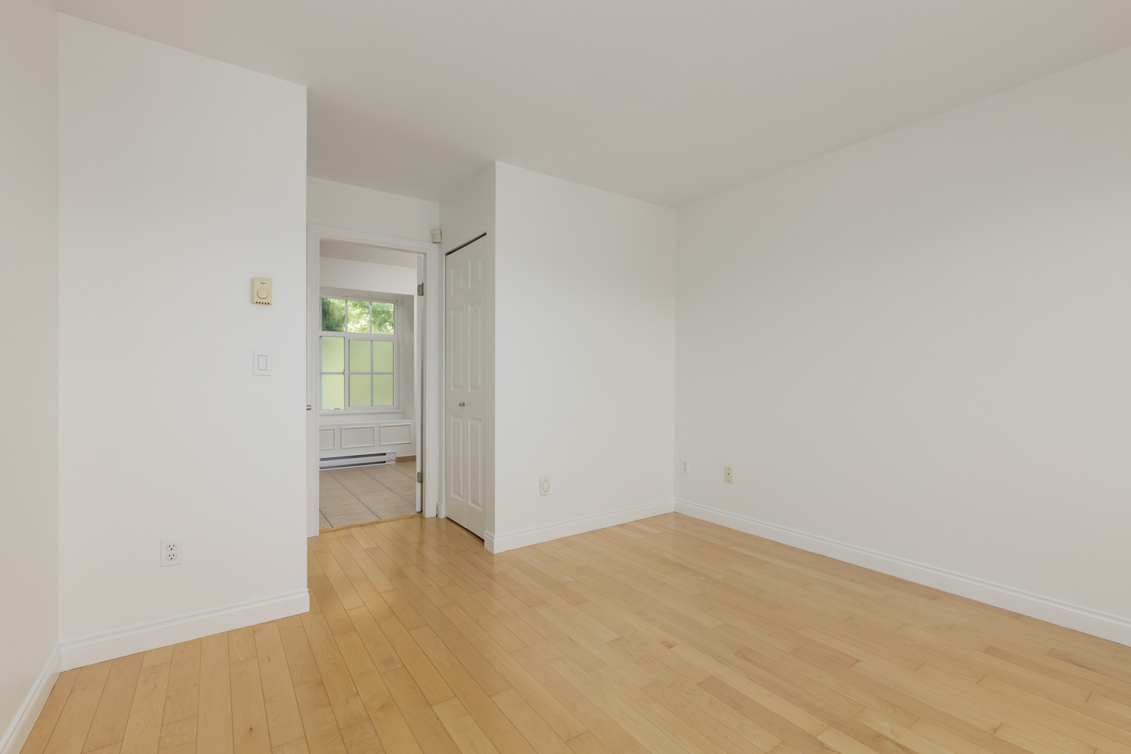 Empty room with light-colored hardwood floor, white walls, and a closet with a white door. An open doorway leads to another room with a window and beige tile floor.