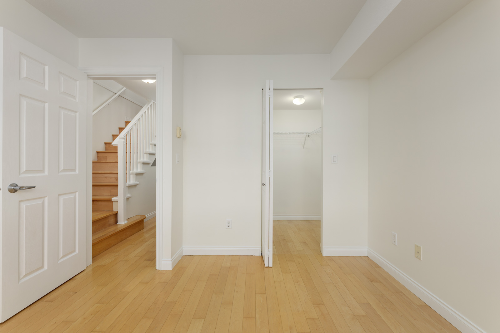 A room with light wood flooring, white walls, an open closet, an open door leading to stairs, and visible electrical outlets.