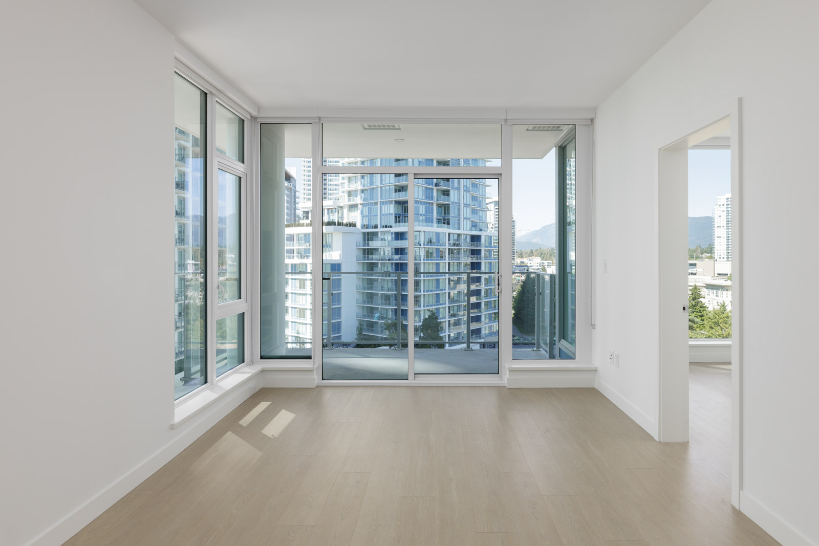 Empty modern apartment room with light wood floors, large floor-to-ceiling windows, and glass doors leading to a balcony with city buildings visible outside.