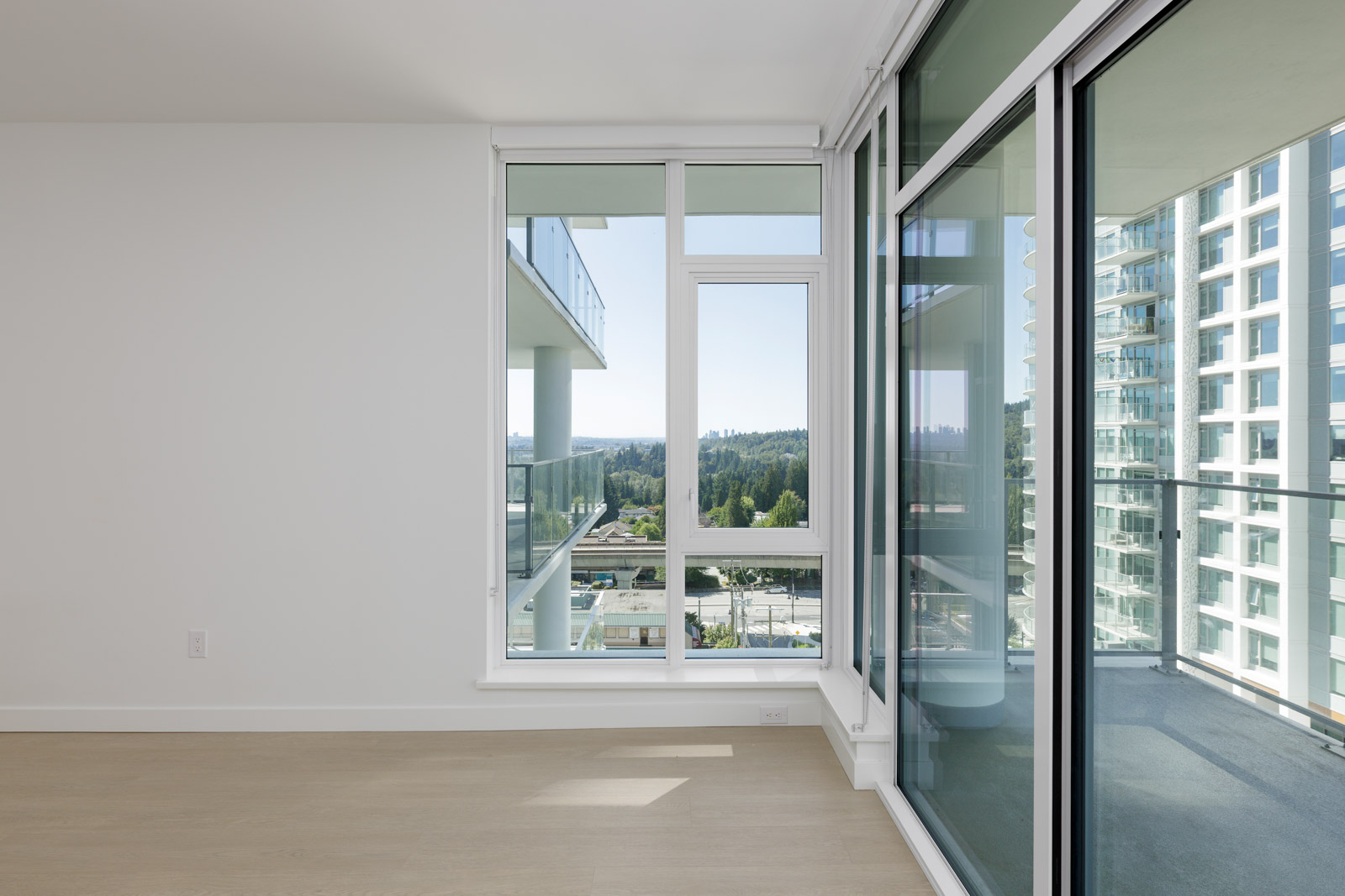 Empty modern apartment with large windows and glass doors opening to a balcony, offering a view of buildings, trees, and distant hills.