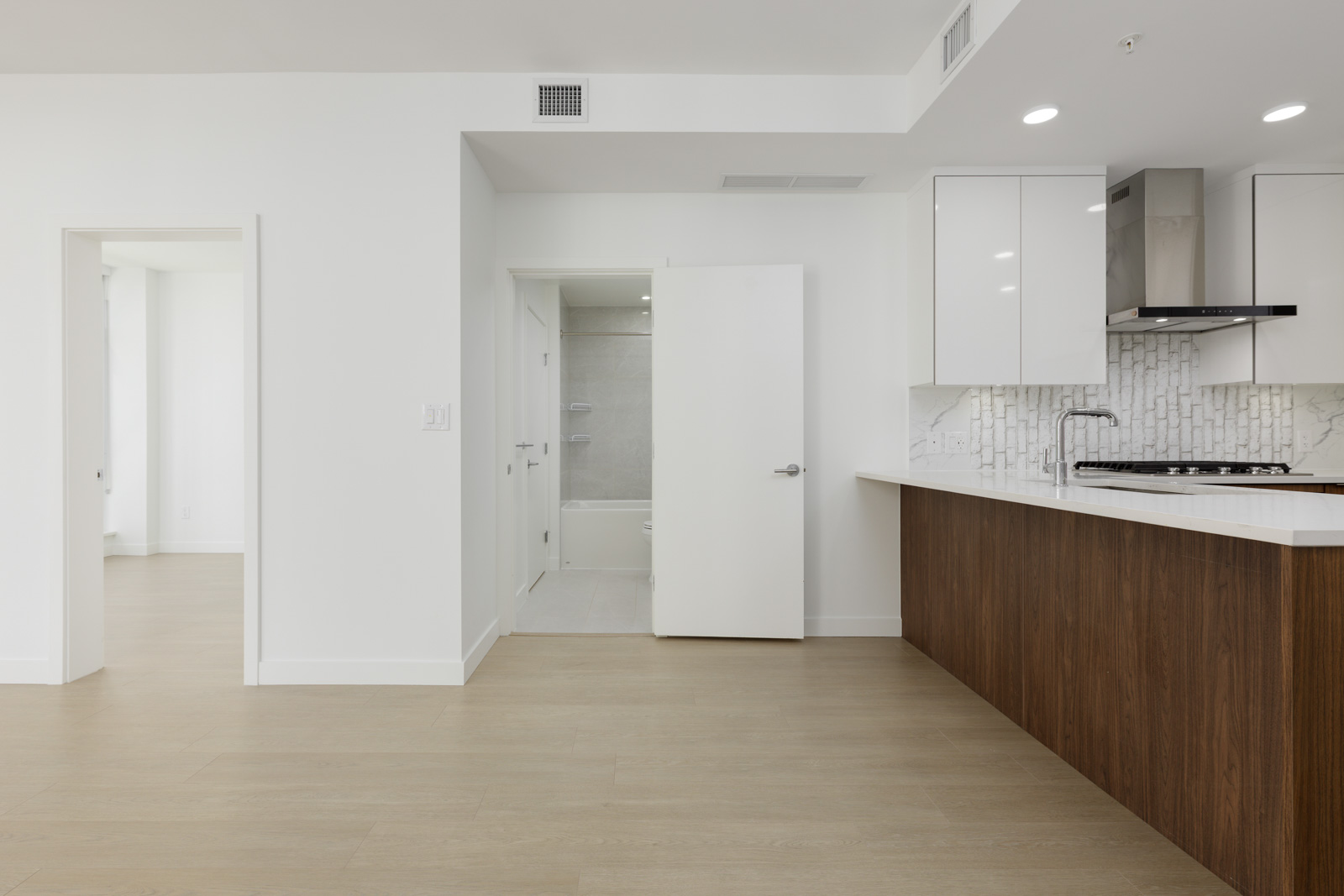 Modern, unfurnished apartment kitchen with white cabinets, wood lower cabinets, and backsplash; open door leads to bathroom and adjacent room visible on the left.