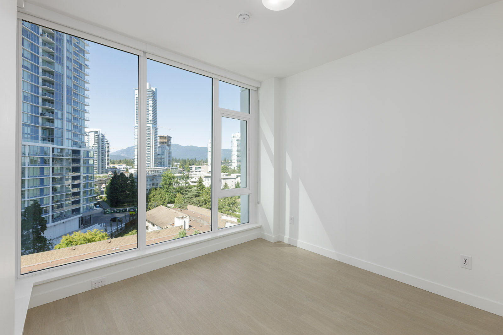 Empty room with light wood flooring, white walls, and large floor-to-ceiling windows showing a cityscape with tall buildings and trees outside.