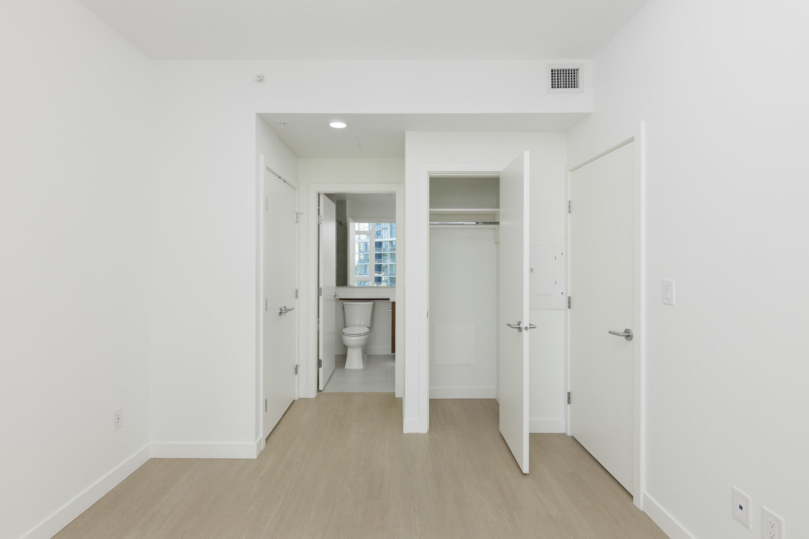A modern, empty bedroom with light wood flooring, white walls, an open closet, and a view into a bathroom with a toilet visible.
