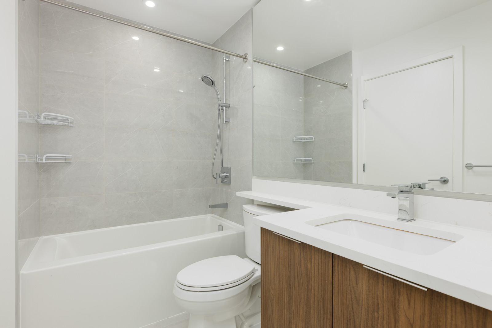 Modern bathroom with light gray tile, white bathtub-shower combo, wall-mounted showerhead, white toilet, and wood vanity with integrated sink and large mirror.