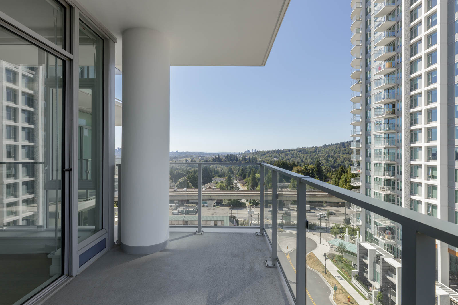 A modern balcony with glass railings overlooks a cityscape, nearby high-rise buildings, a road, and distant trees under a clear sky.