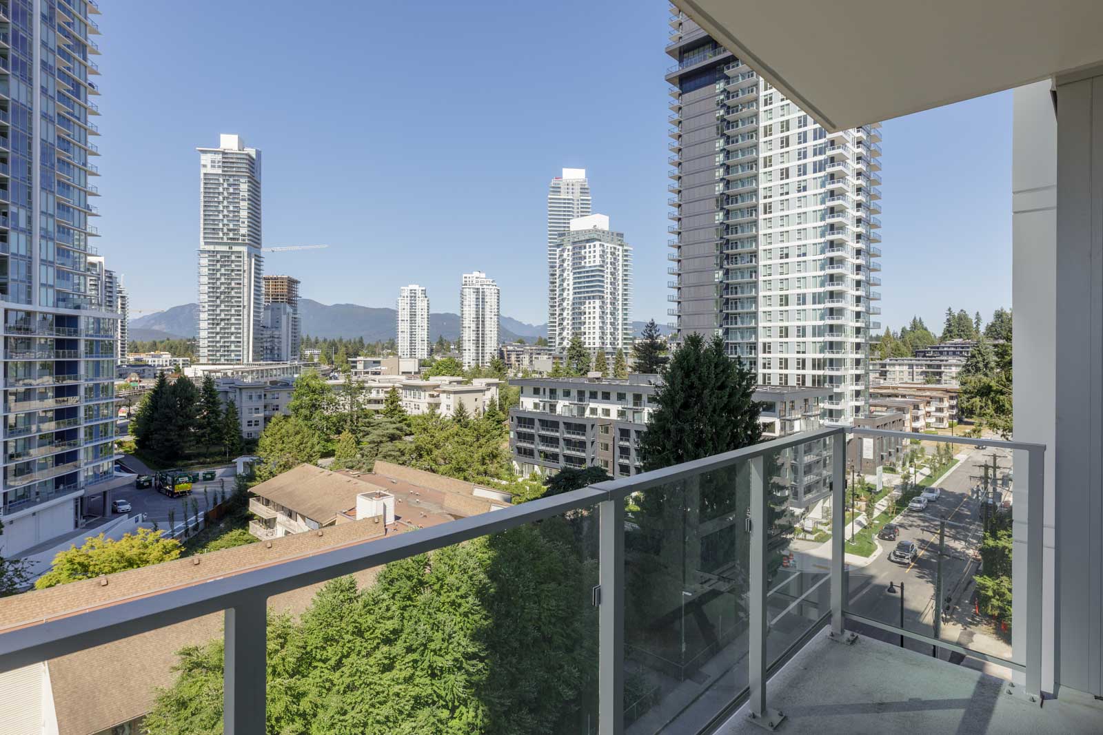 View from a balcony showing glass railings, several high-rise apartment buildings, trees, and mountains in the distance under a clear blue sky.