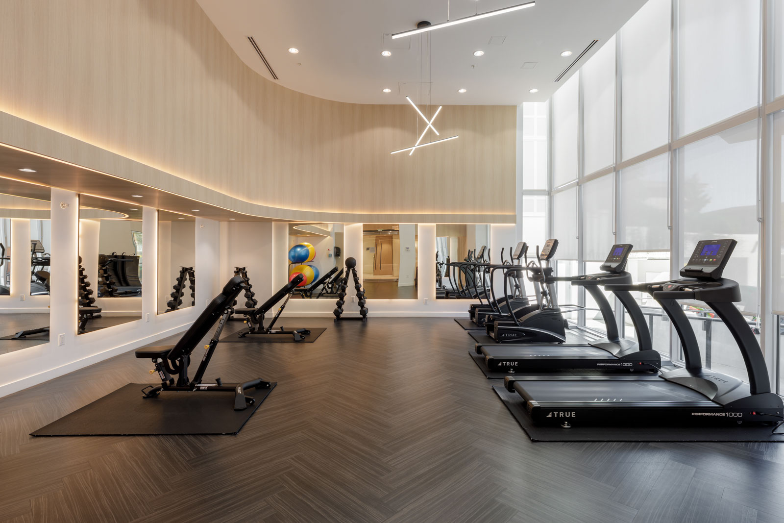 Modern gym with treadmills, resistance machines, large mirrors, wood panel walls, dark herringbone flooring, and floor-to-ceiling windows allowing natural light inside.