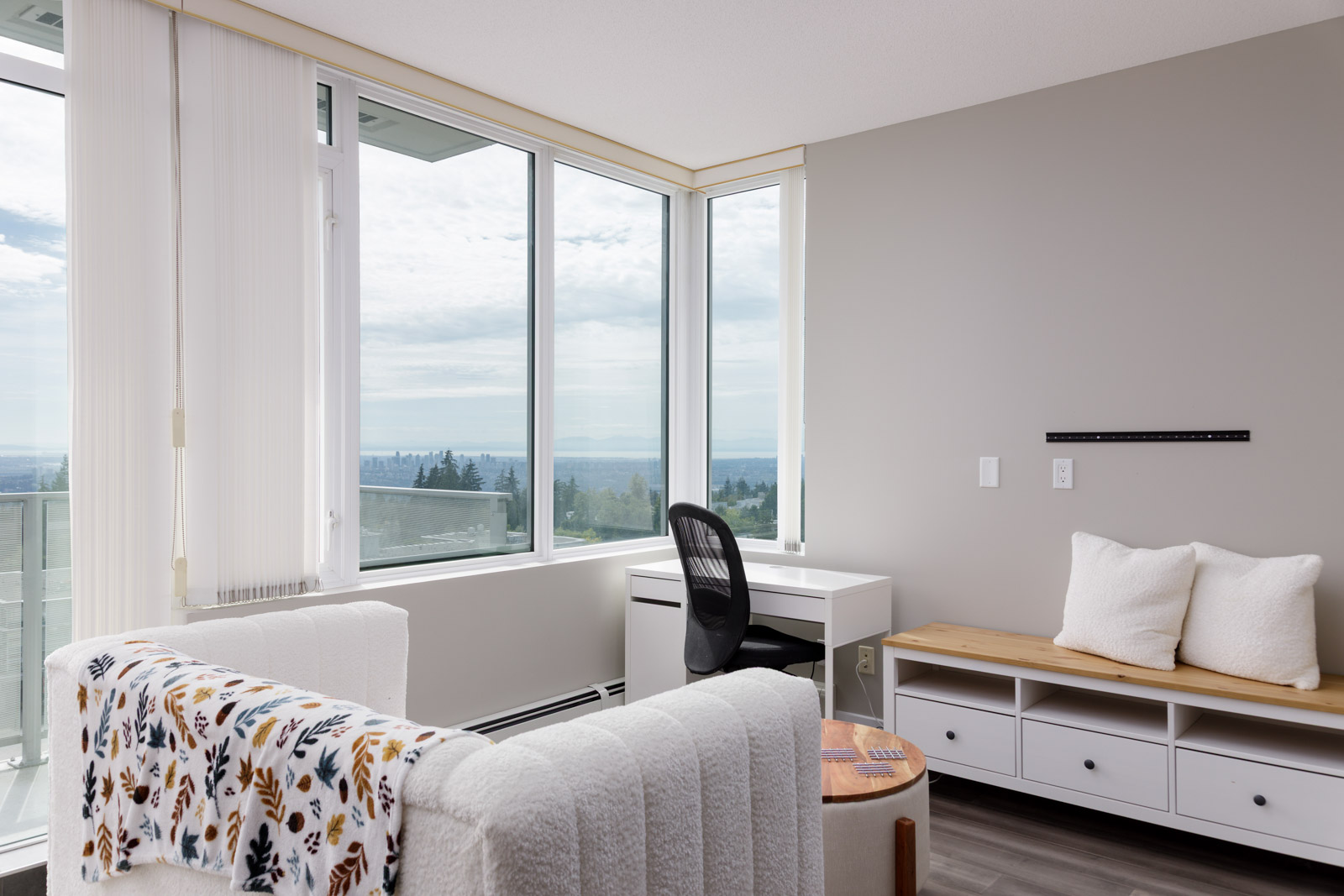 Bright, modern room with large windows, a white desk and chair, cushioned bench, and a patterned blanket on a sofa. Cityscape and sky visible outside.