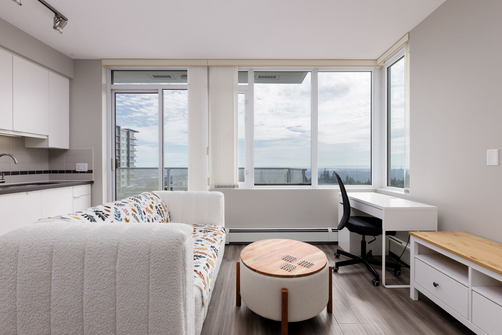 A modern, bright living area with a sofa, round coffee table, white desk and chair, large windows, and a view of buildings and the ocean outside.