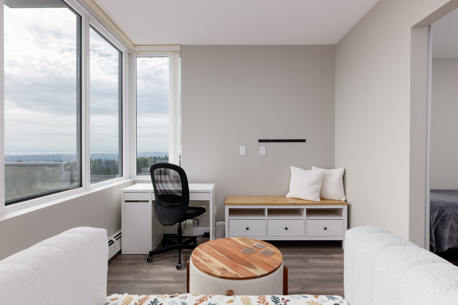 A bright room with a large window, a white desk and office chair, a wooden bench with storage, a round coffee table, and part of a bed visible through an open doorway.