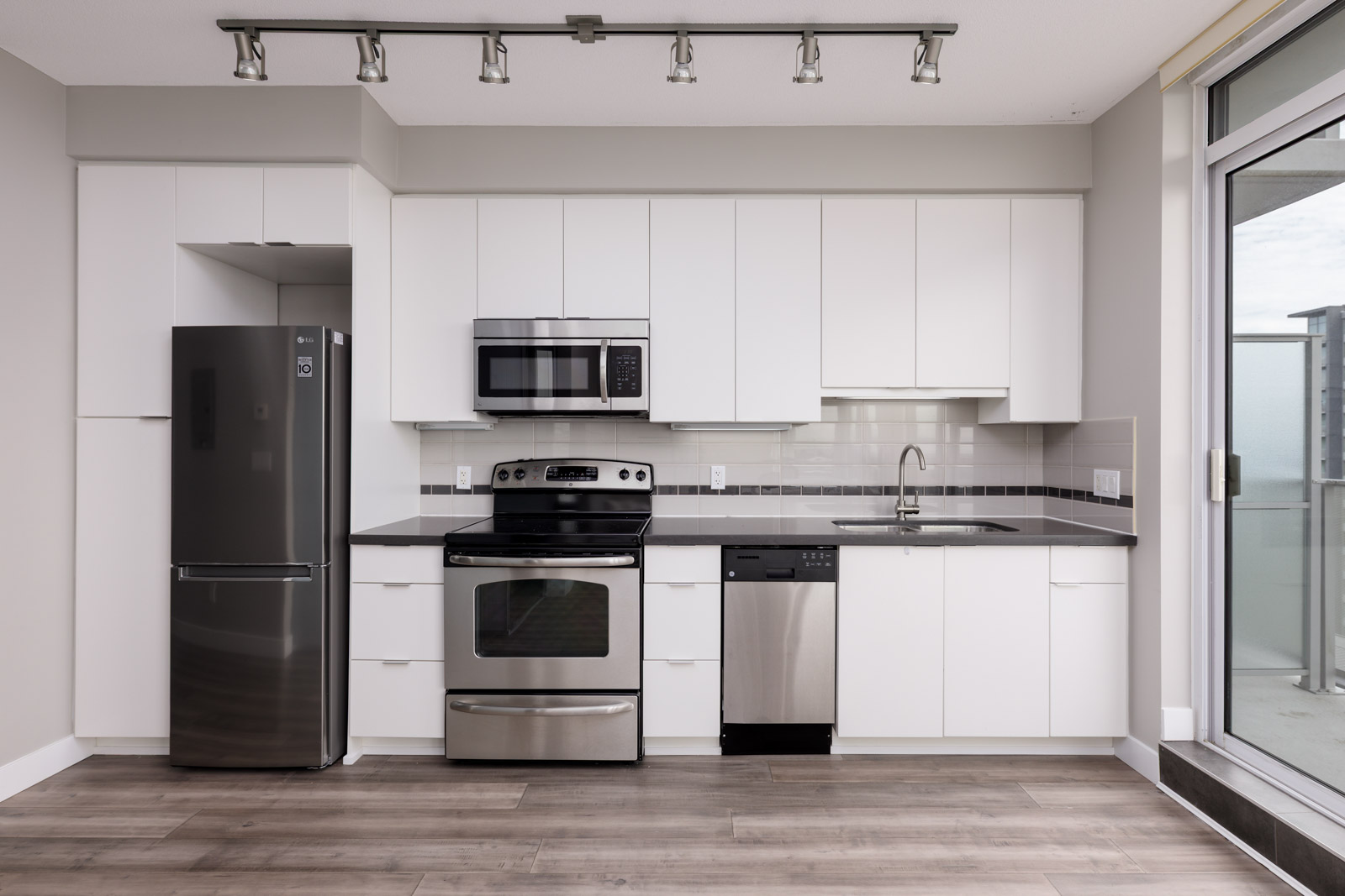 Modern kitchen with white cabinets, stainless steel appliances including fridge, oven, microwave, and dishwasher, light wood flooring, and a sliding glass door to a balcony.