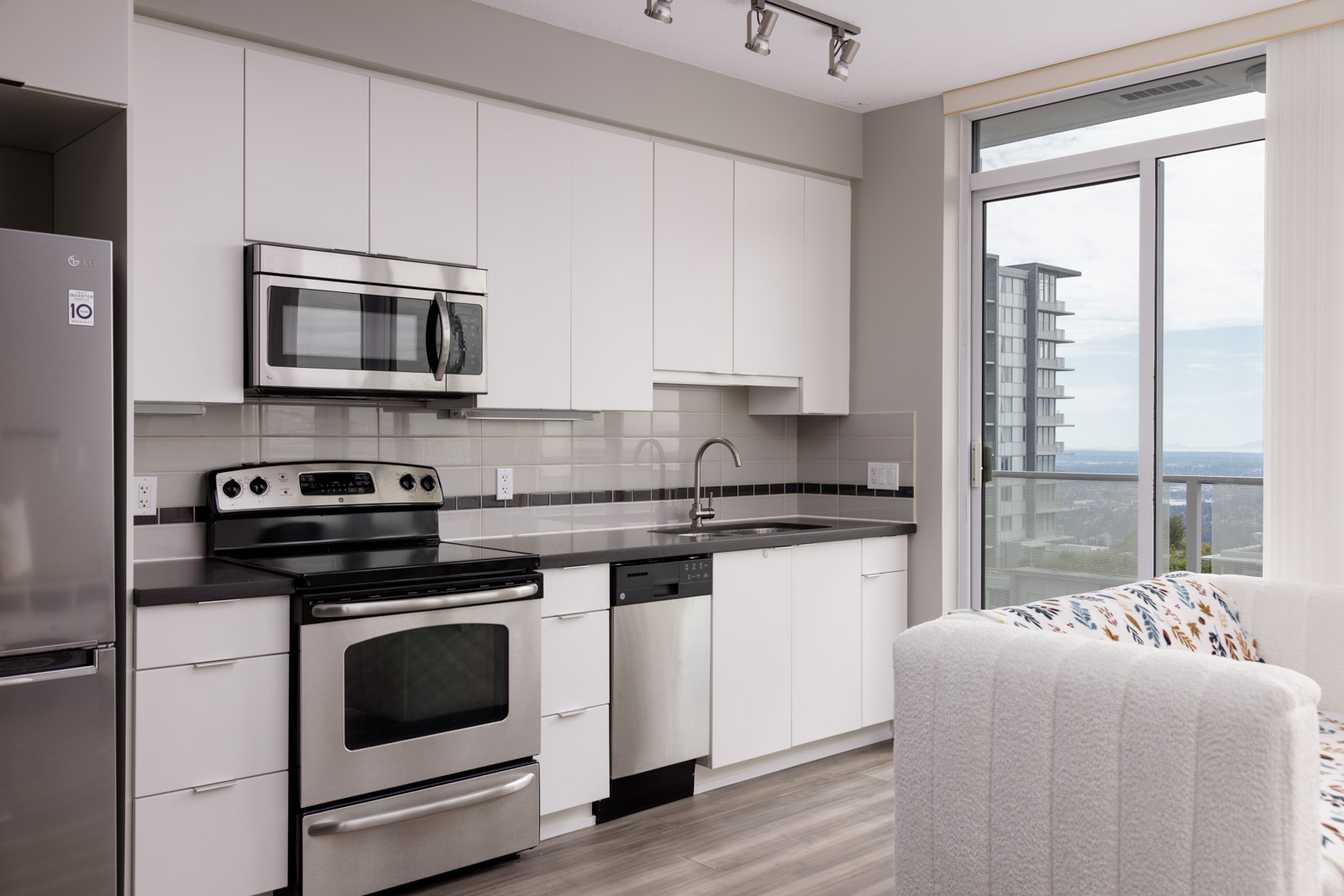Modern apartment kitchen with white cabinets, stainless steel appliances, a sink, and a dishwasher; large window with city view and part of a white sofa visible.