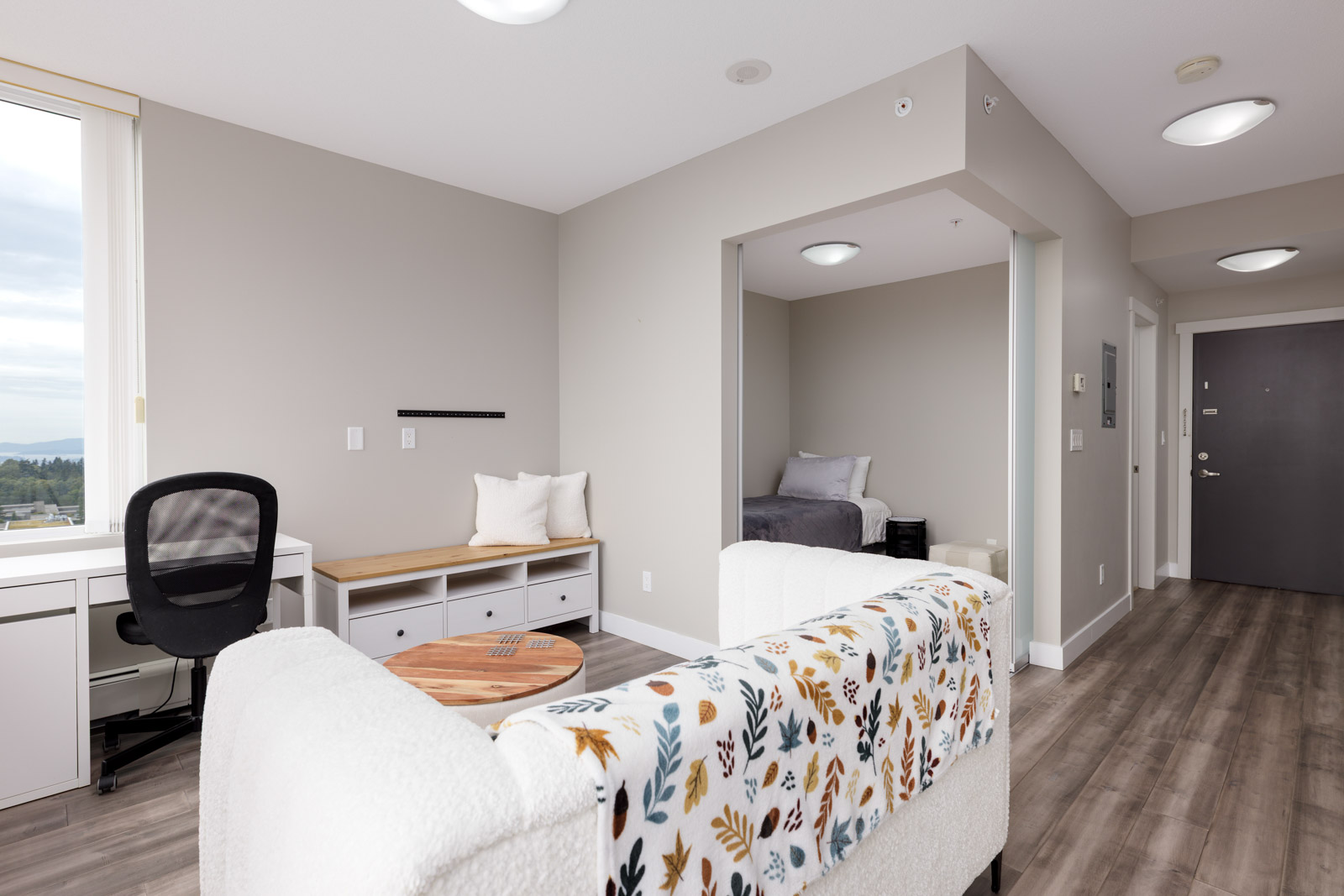 A modern studio apartment with a bed alcove, desk, seating area, and large window; neutral colors and wood flooring throughout.