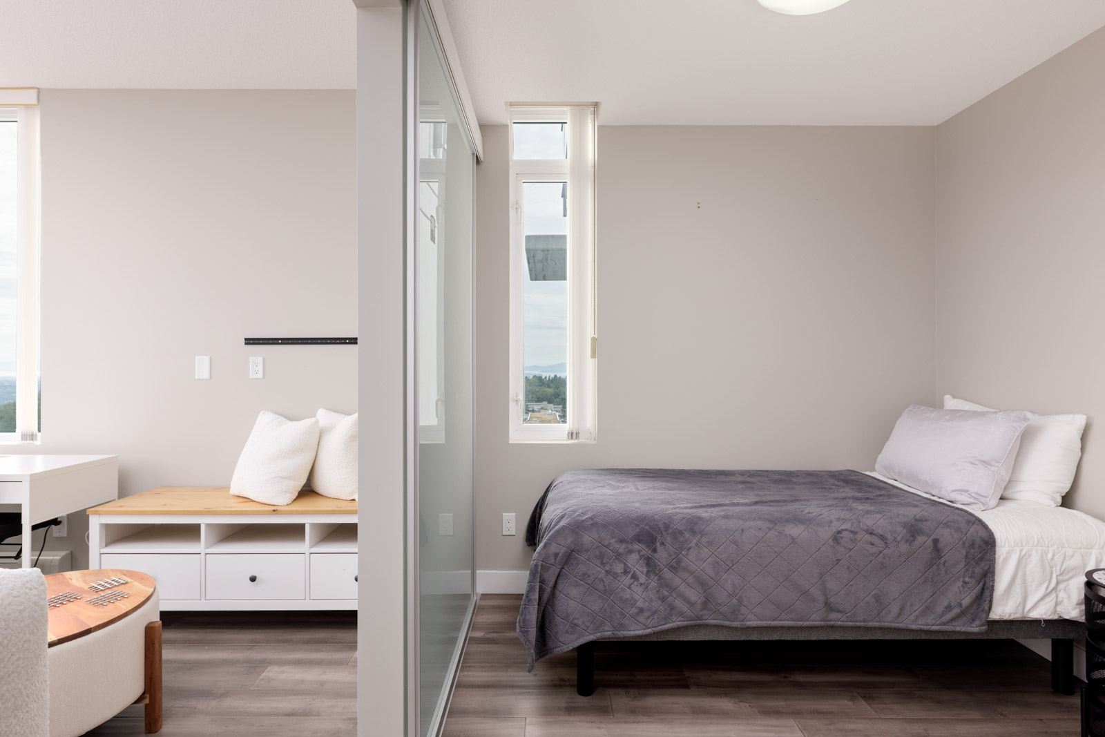 A minimalist bedroom with a bed, gray quilt, white pillows, a desk, bench, and large windows, separated by a glass partition.