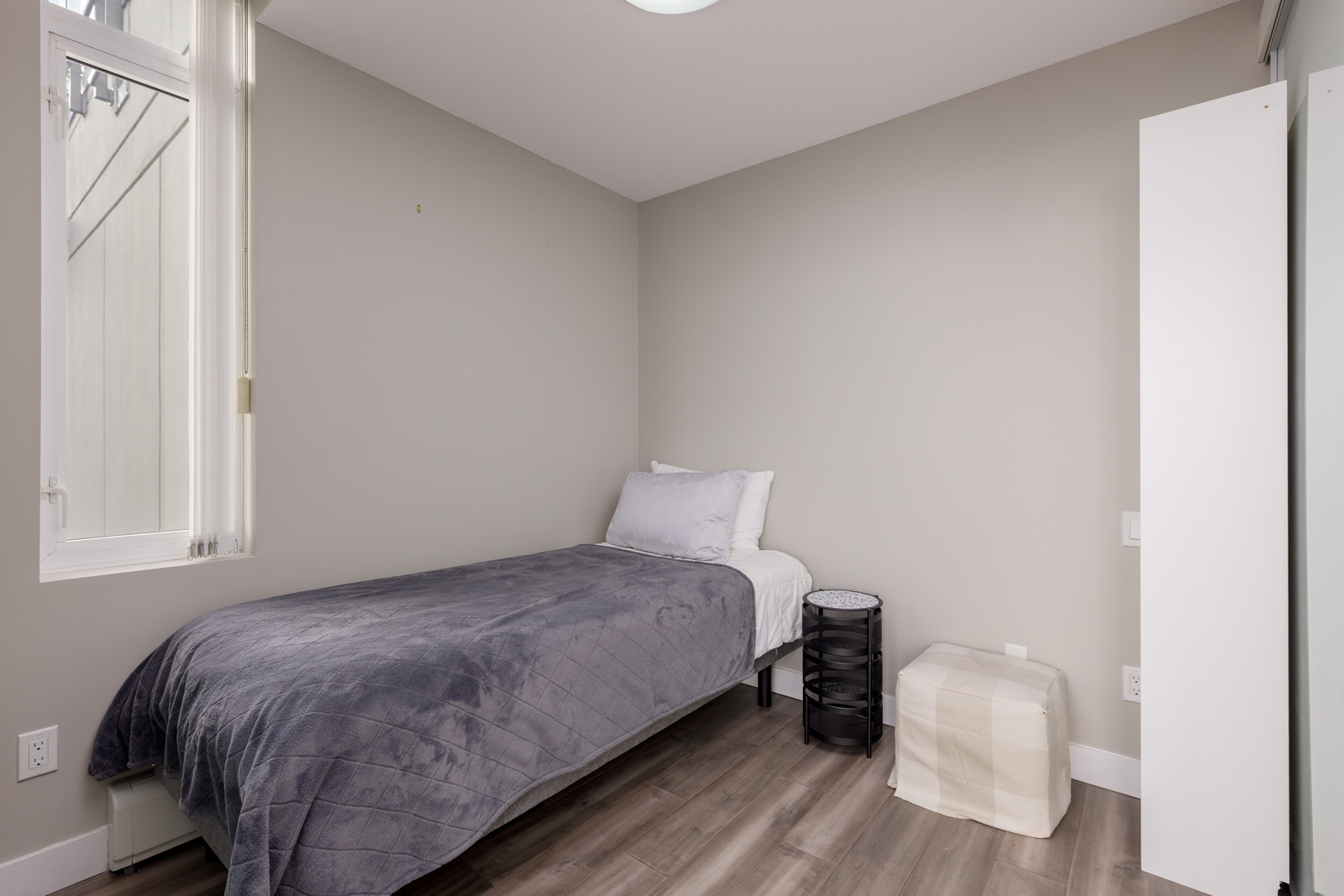 A small bedroom with a single bed, gray blanket, white pillows, a round black nightstand, a beige pouf, and a window letting in natural light.