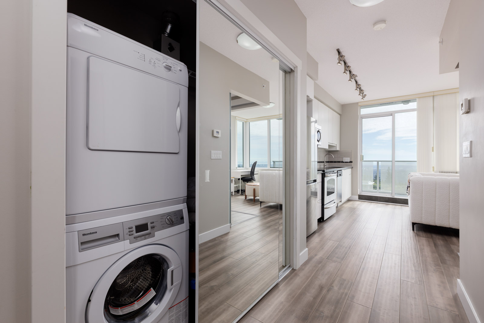 Stacked washer and dryer in a laundry closet next to a modern kitchen and living area with large windows and a balcony view.