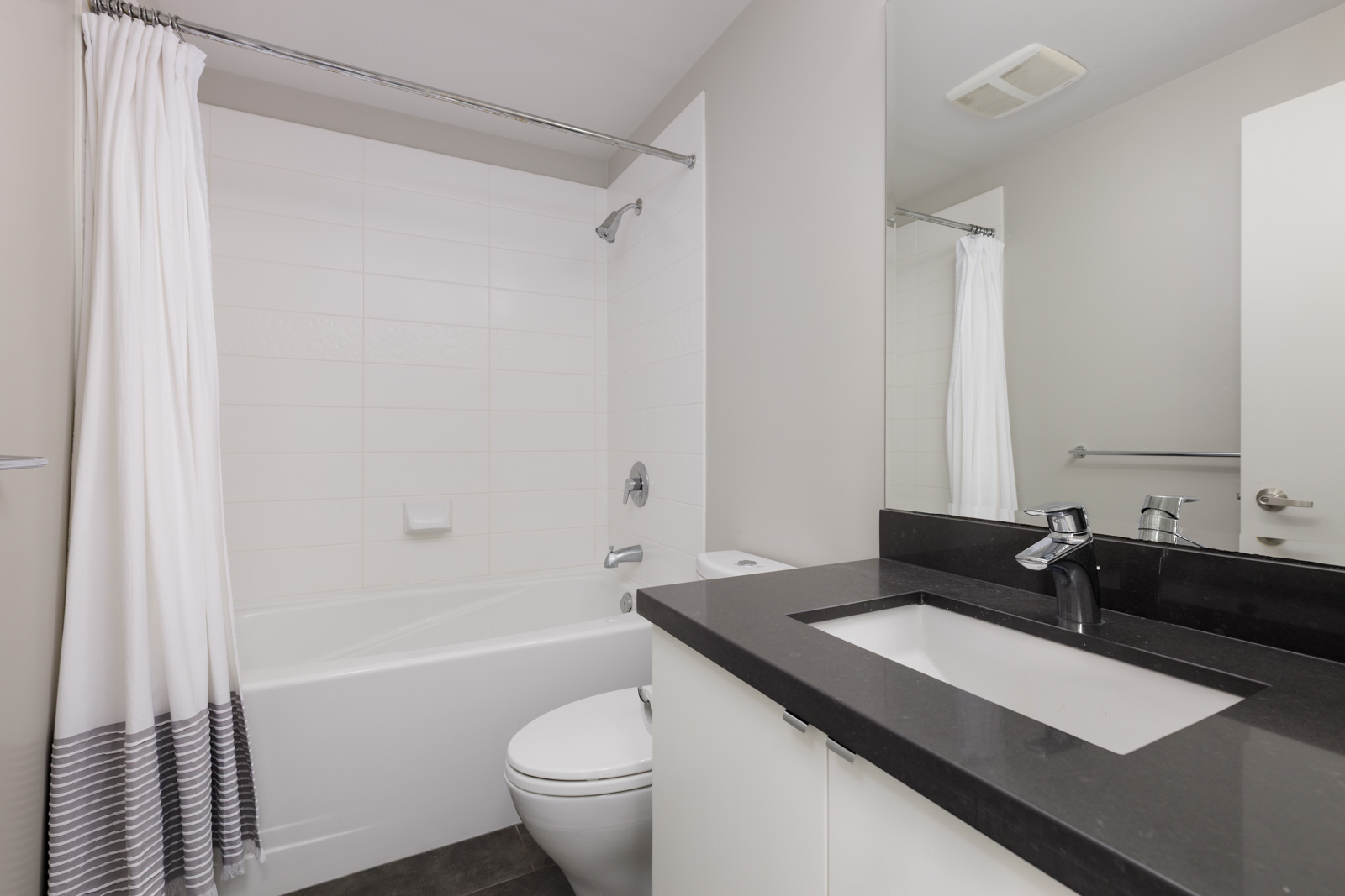 Modern bathroom with a white bathtub and shower, white curtain, toilet, black countertop, and a white sink with a large mirror above.