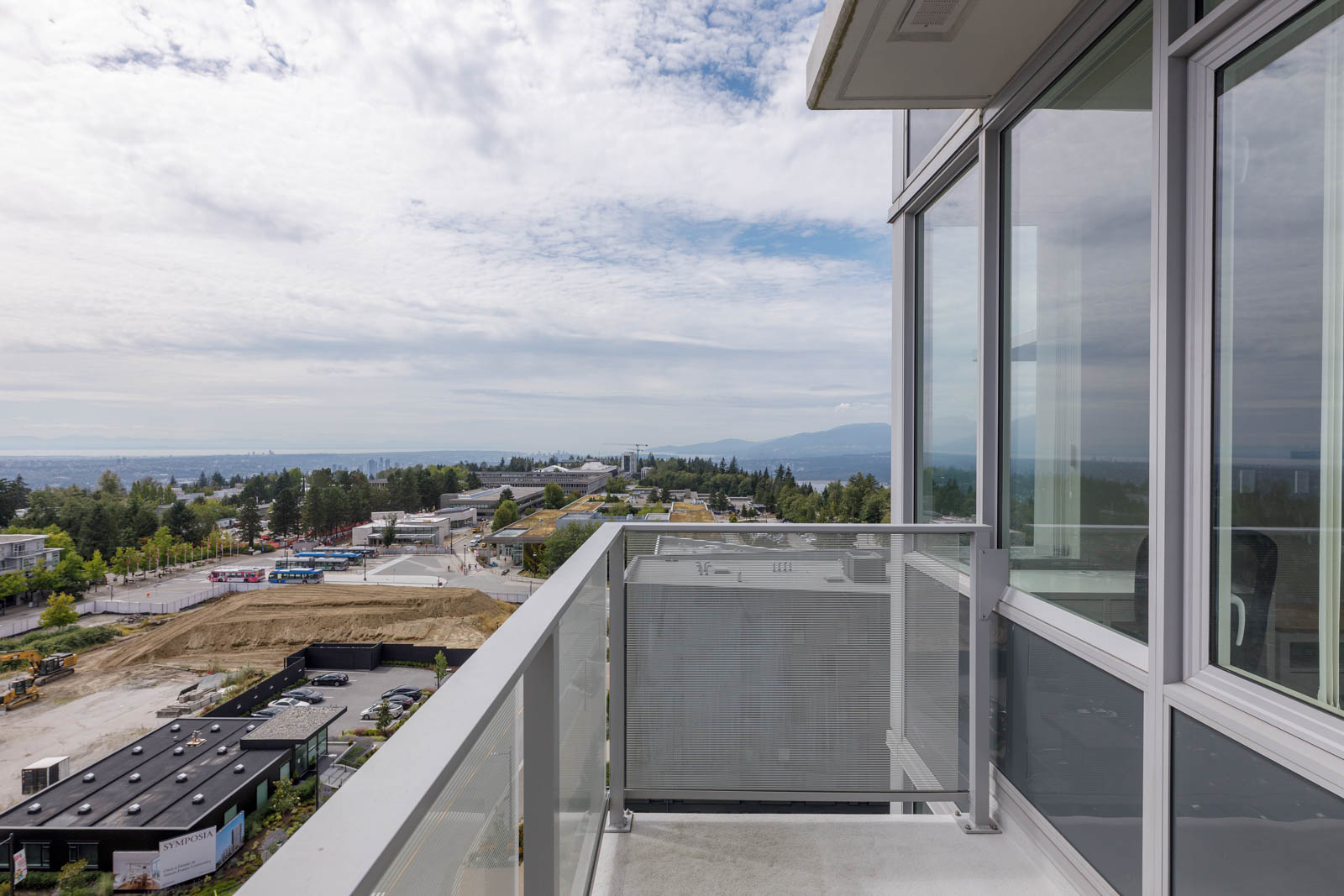 View from a modern balcony overlooking a suburban area with trees, buildings, a construction site, and mountains in the background under a partly cloudy sky.