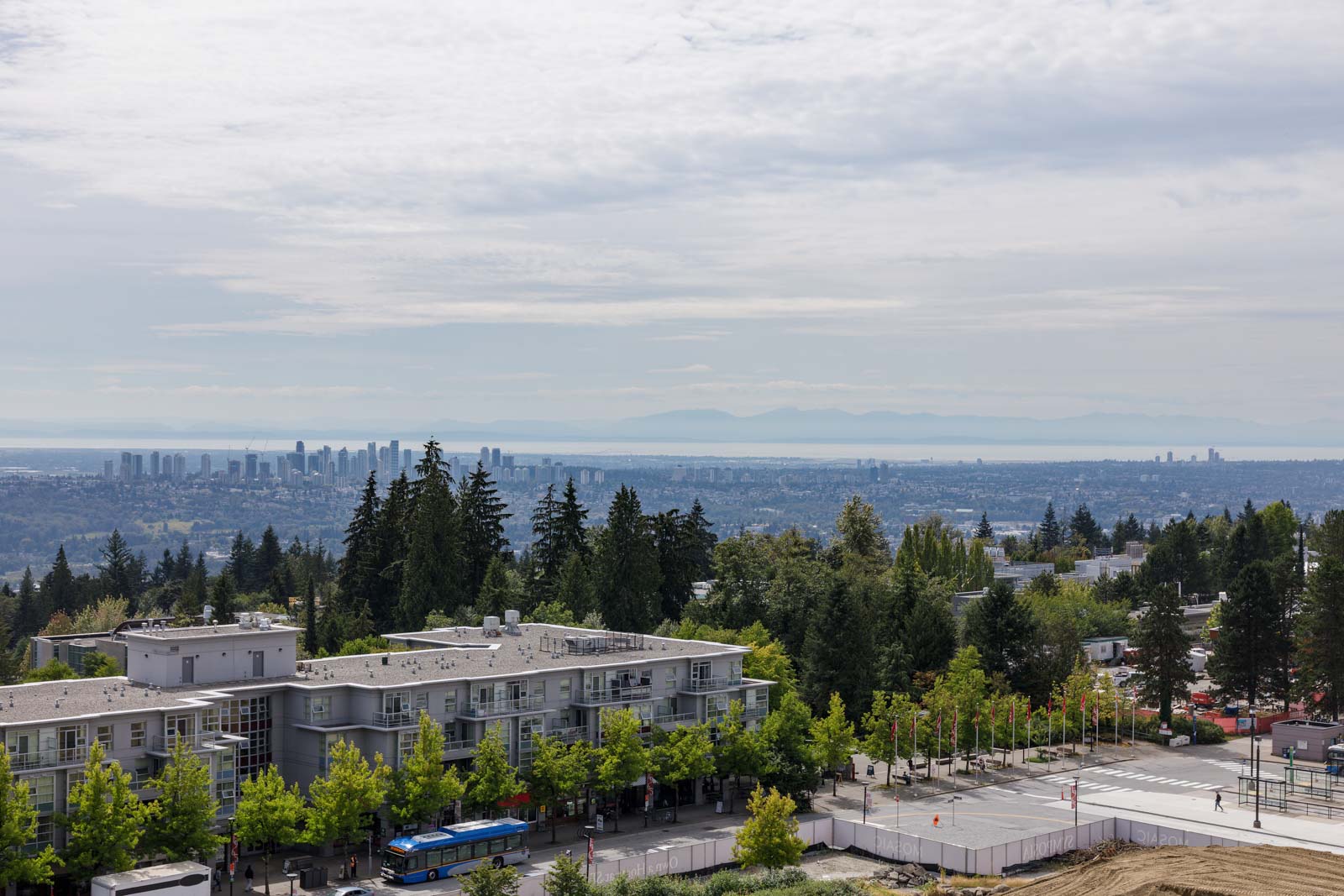 View of a suburban area with mid-rise buildings, tree-lined streets, and a distant city skyline under a cloudy sky.