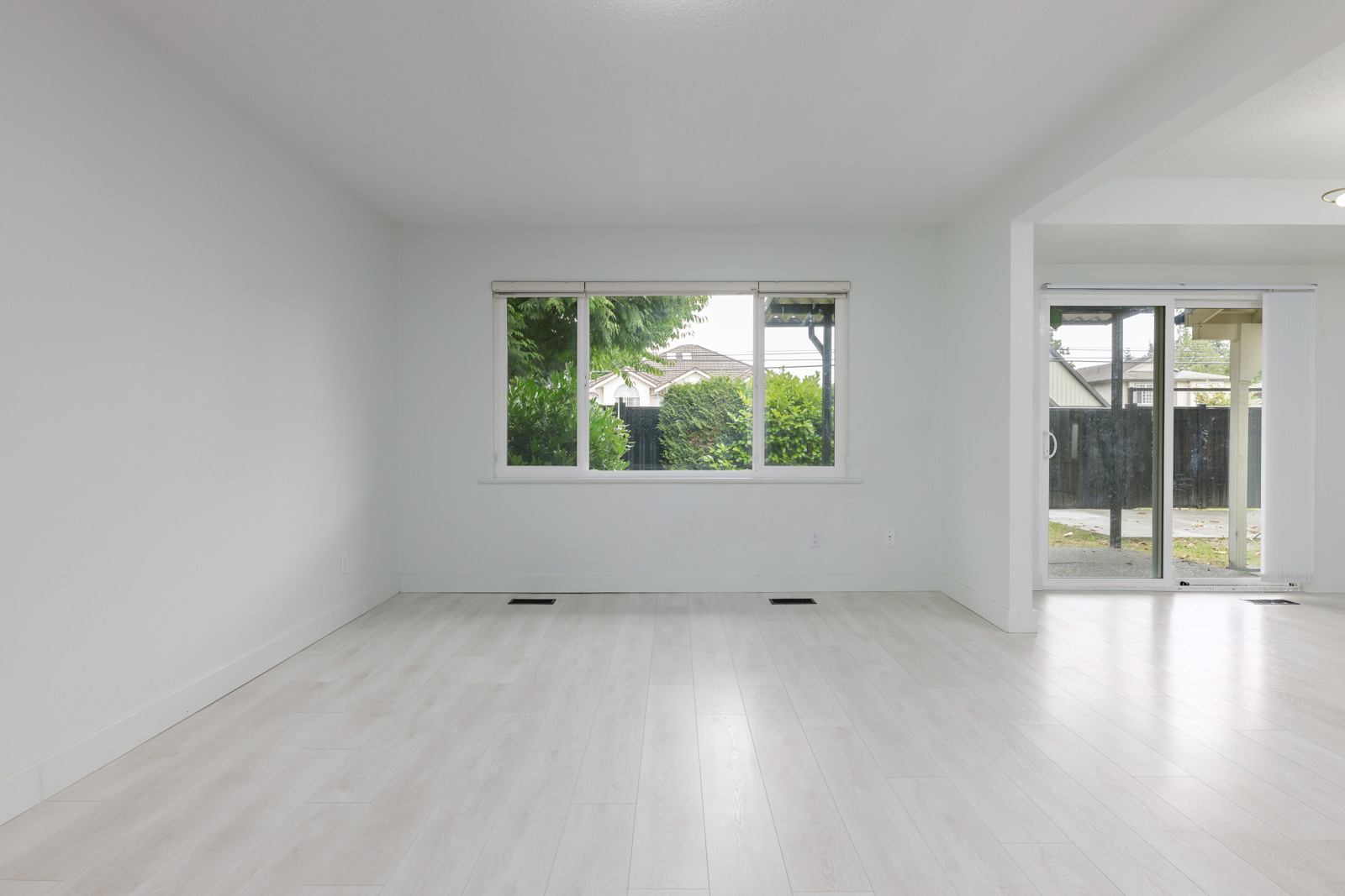 Empty, unfurnished room with white walls, light wood flooring, a large window, and sliding glass doors leading to an outdoor area.