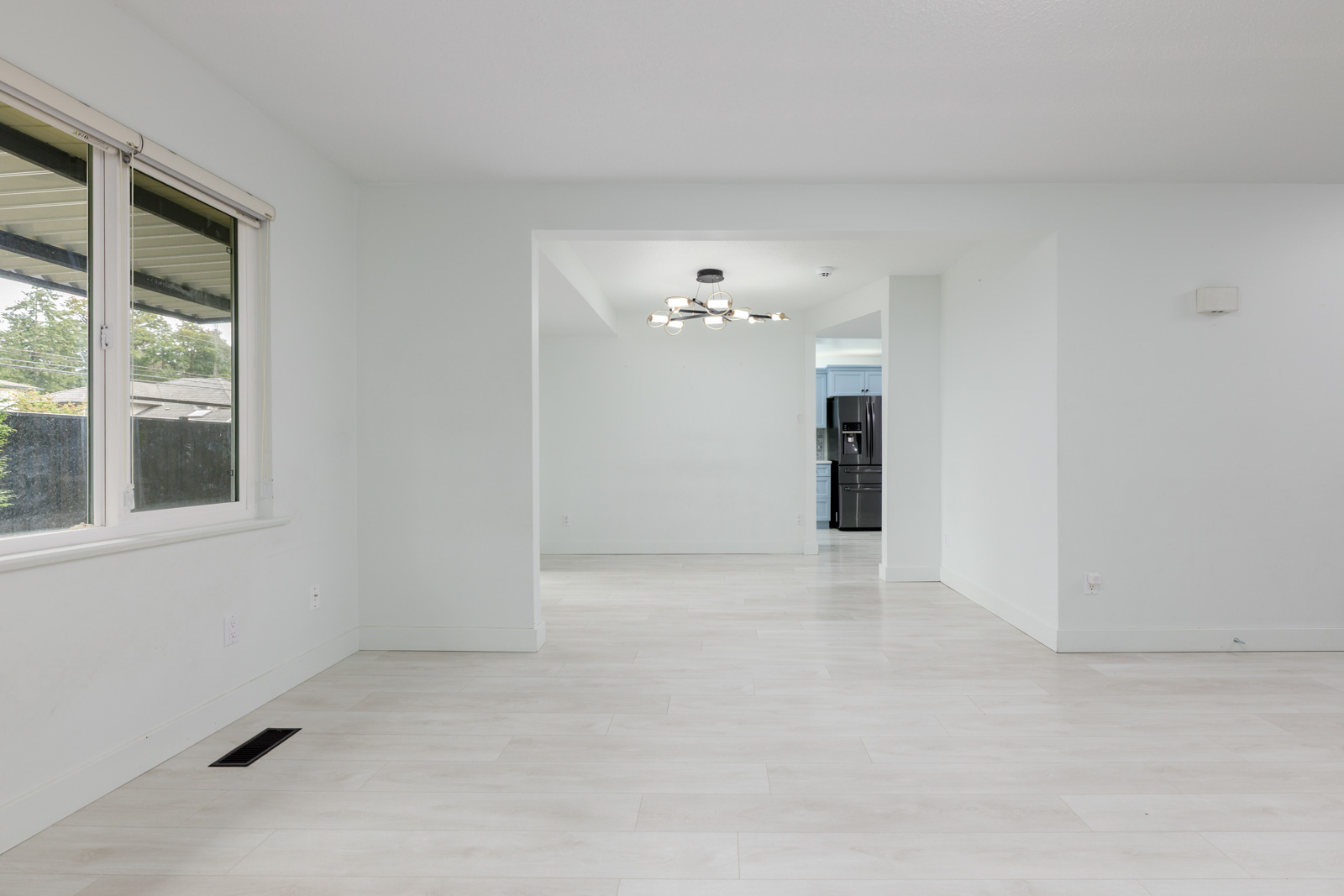 Empty, unfurnished open-plan room with white walls and light-colored floors, large front window on the left, and a view into the adjoining kitchen area with a modern light fixture.