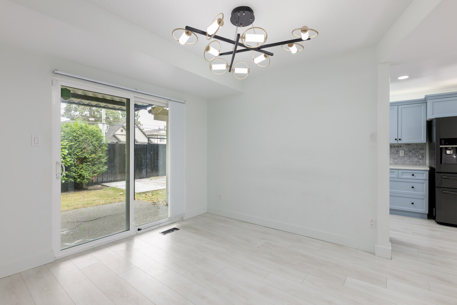 Bright, empty room with light wood flooring, modern black chandelier, large sliding glass doors to a patio, and partial view of a blue kitchen with black appliances.