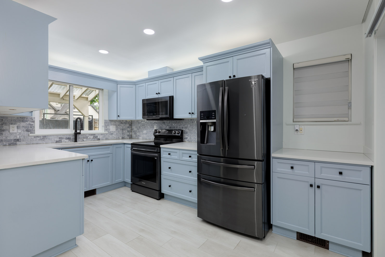 Modern kitchen with light blue cabinets, black appliances, white countertops, tile backsplash, and a window above the sink.