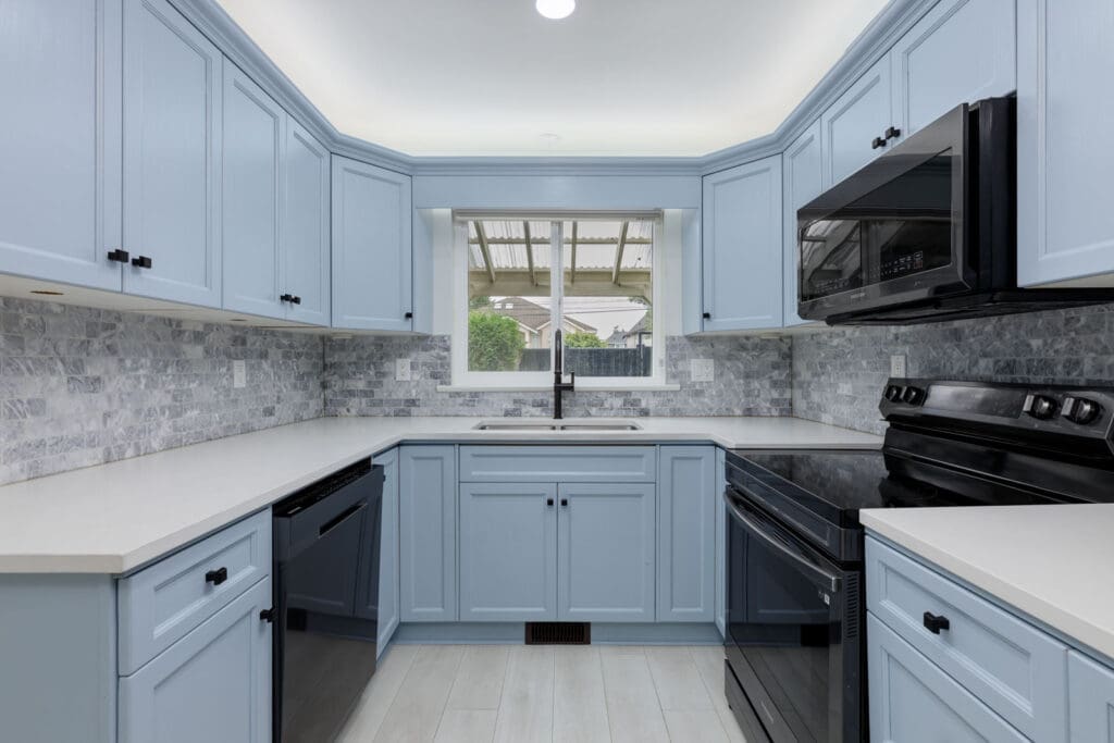 Modern kitchen with light blue cabinets, white countertops, black appliances, gray tile backsplash, and a window above the sink.