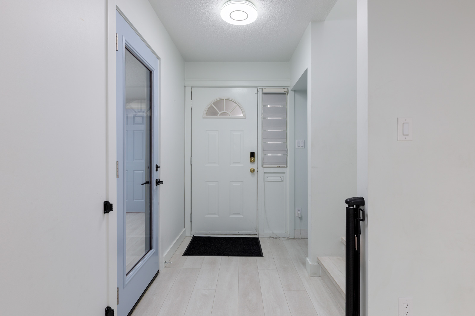 A white entryway with a main door, a side window, a glass door to the left, light wood flooring, and a small black doormat in front of the entrance.