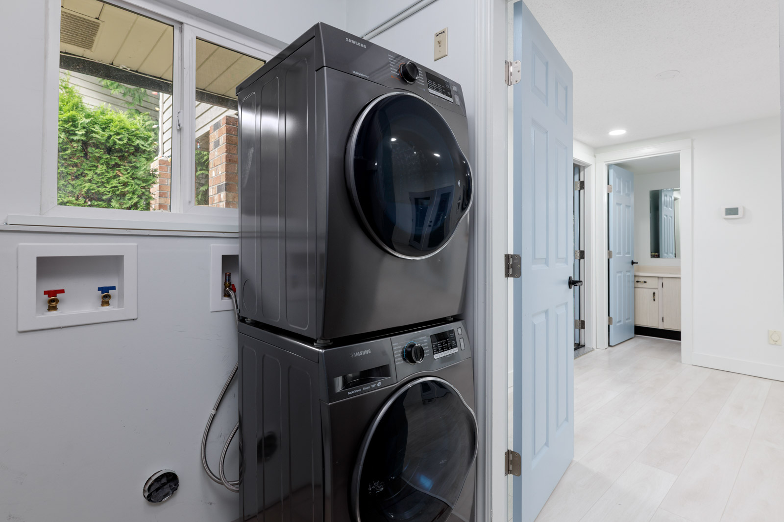 A laundry room with a stacked black washer and dryer, open utility hookups on the wall, and a view into a bathroom with a vanity and mirror.