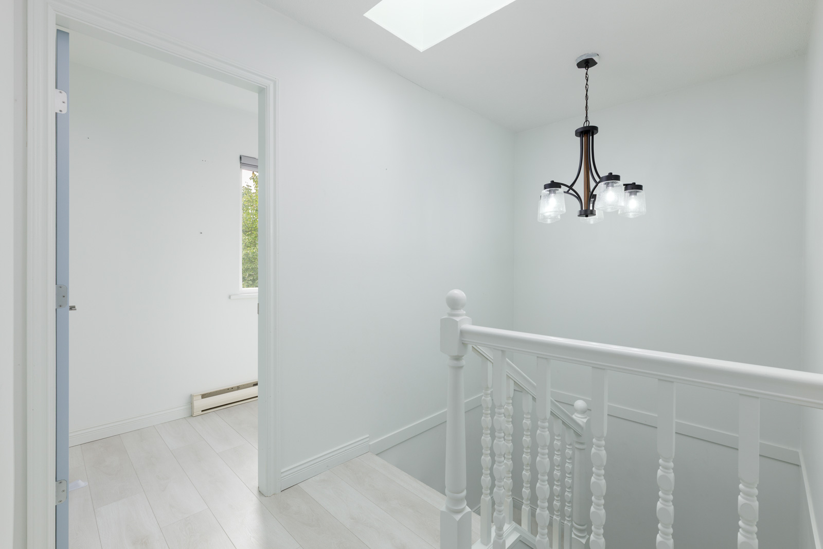 A white hallway with a staircase, a modern black chandelier, light wood floors, and a doorway leading to a bright room with a window.