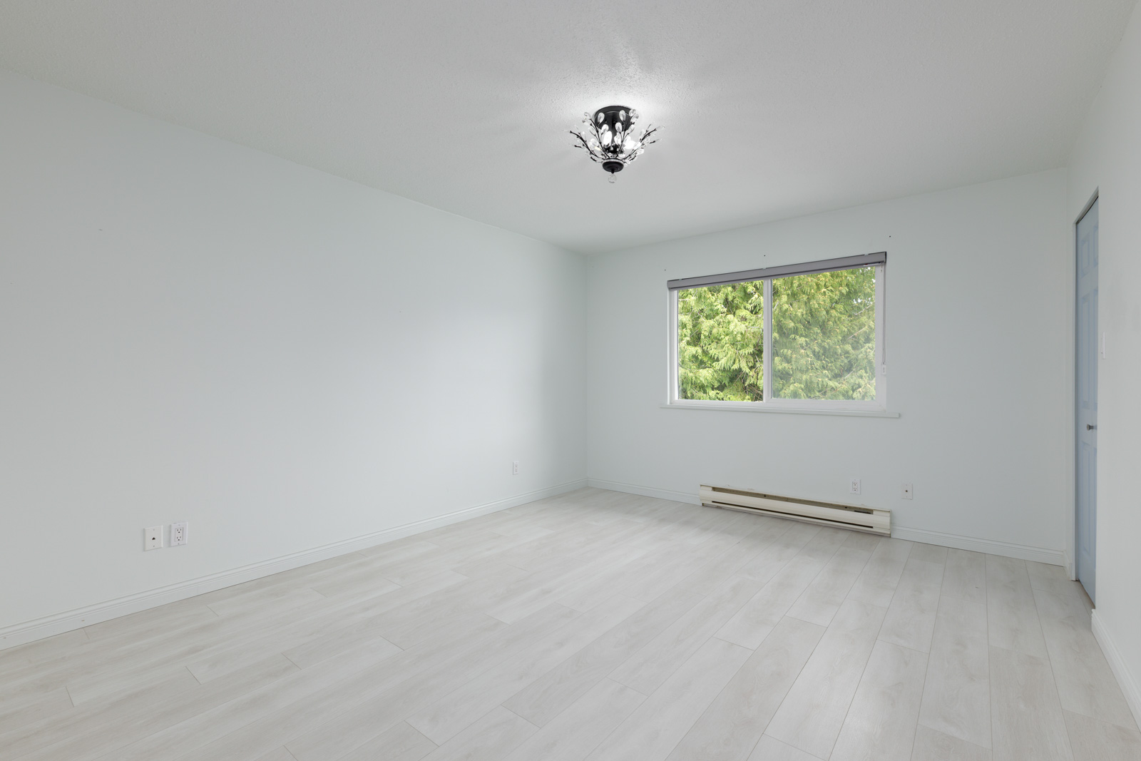Empty room with light-colored walls and flooring, a ceiling light fixture, window with a view of trees, and a blue door on the right.