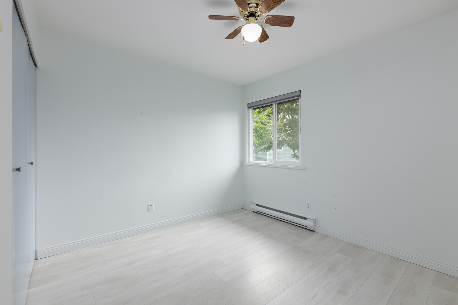 Empty room with light-colored walls and flooring, a ceiling fan with a light, a window with blinds, and a baseboard heater below the window.