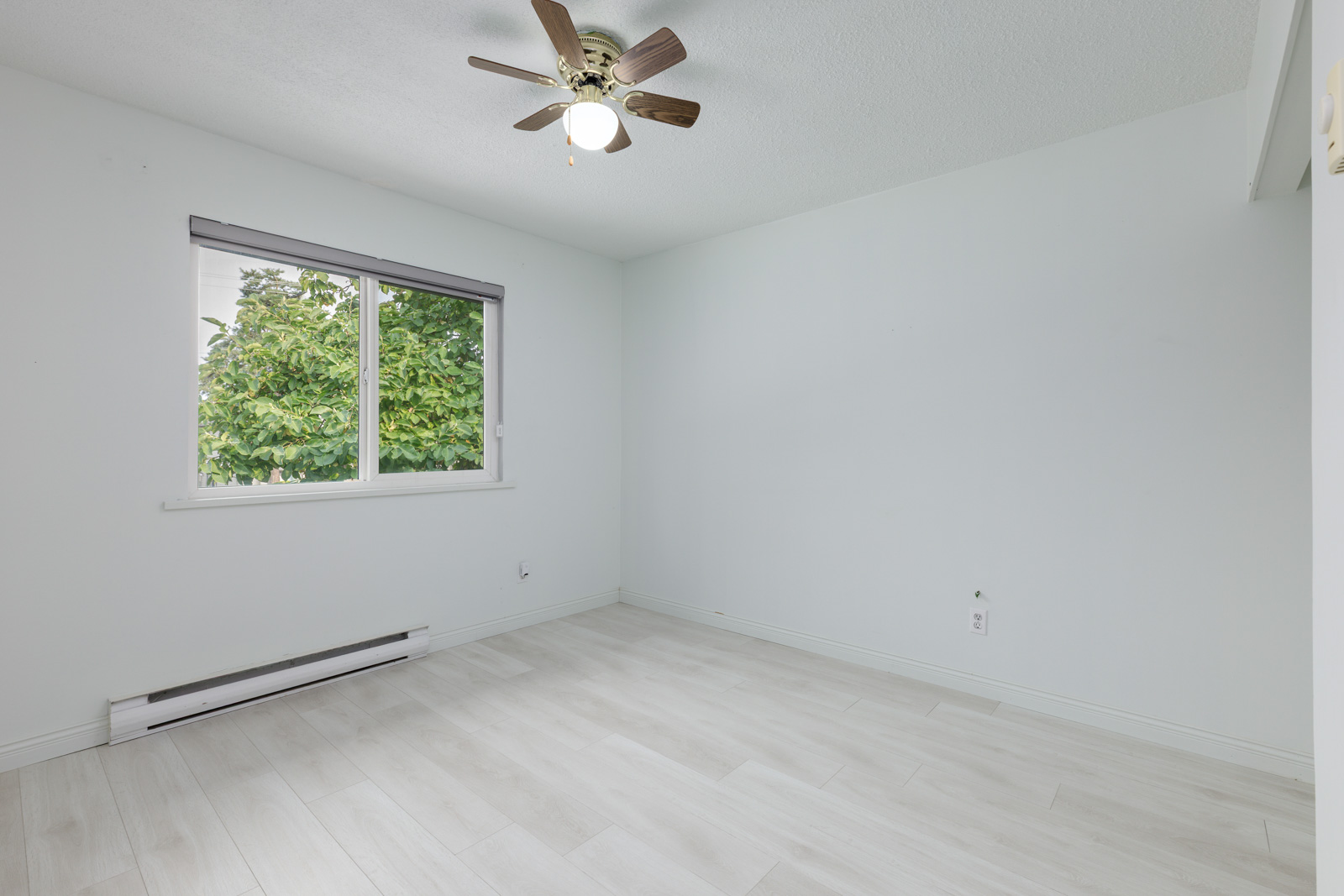 Empty room with light-colored flooring, white walls, a window with a view of trees, ceiling fan with light, and an electric baseboard heater under the window.