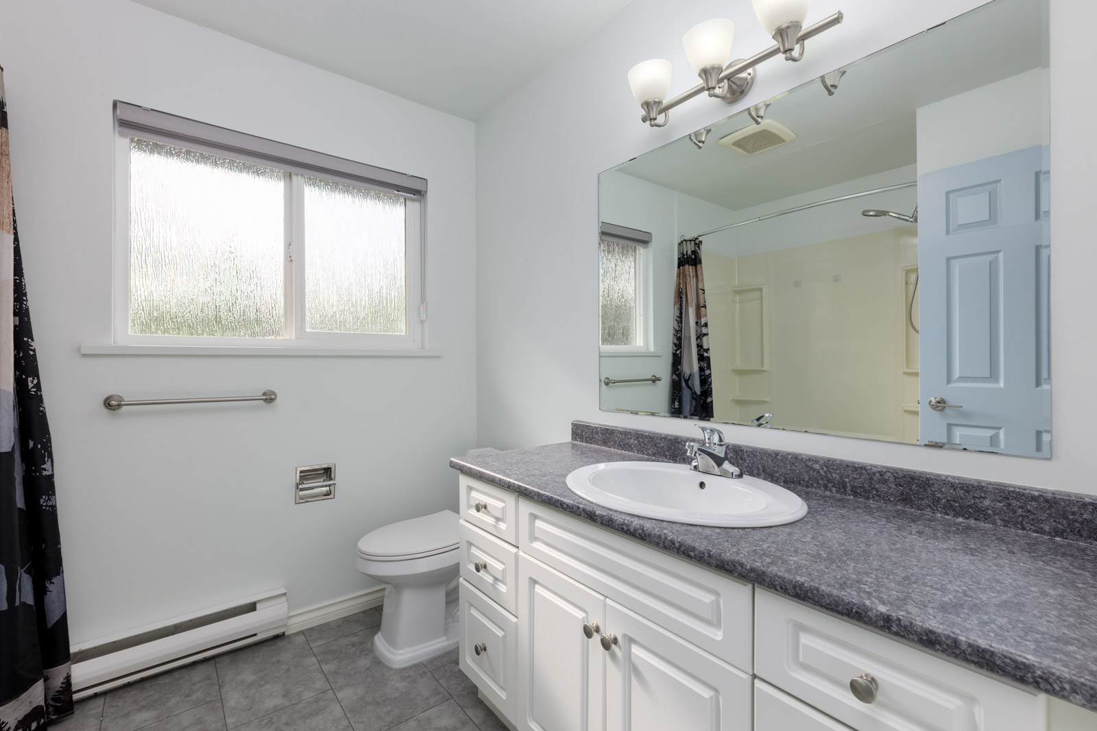 A clean, white bathroom with a large mirror, single sink vanity, toilet, frosted window, and gray countertop and flooring.