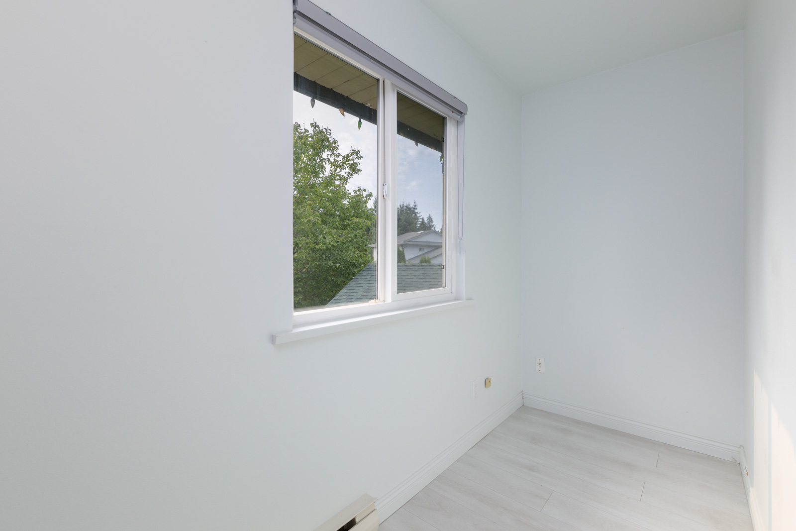 Small, empty room with light-colored walls and flooring, a double window with a partial view of trees and rooftops outside, and a white electrical outlet on the wall.