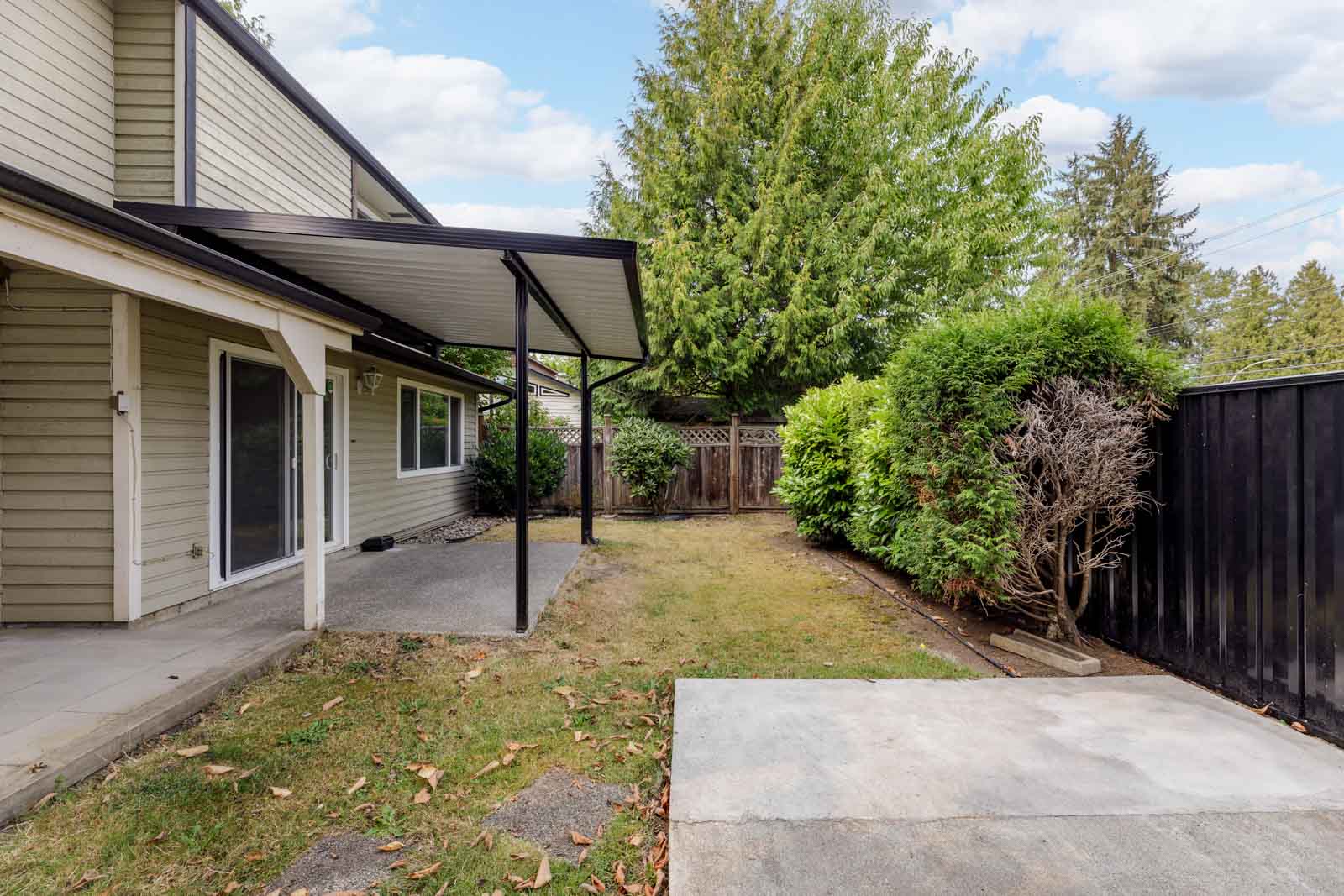 A backyard with a covered patio area, concrete slab, lawn, and surrounding trees and hedges next to a beige house.