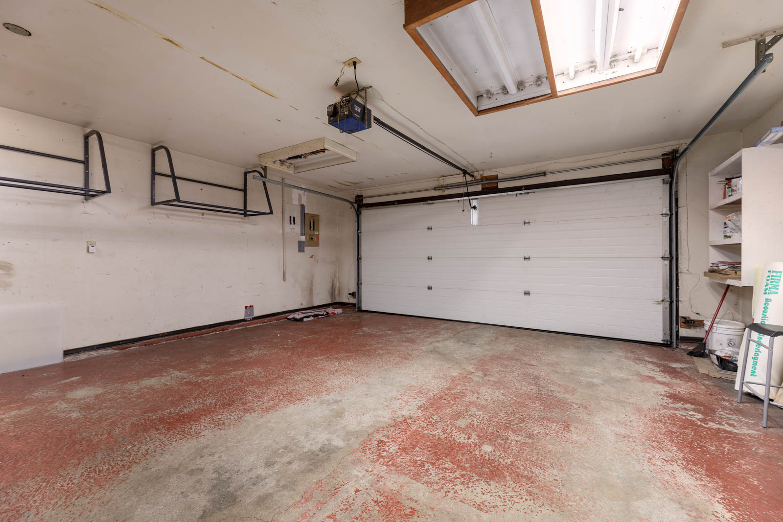 Empty two-car garage with a red-stained, worn concrete floor, white walls, overhead storage racks, shelves on the right, and a closed garage door.