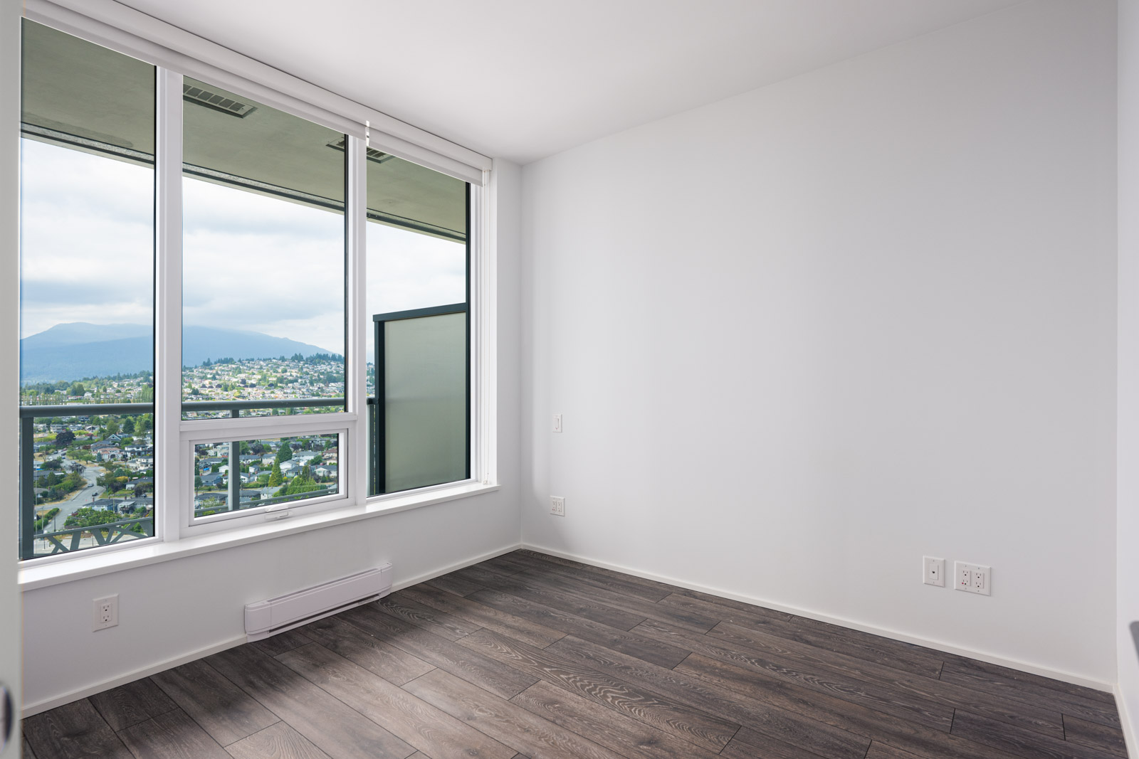Empty room with white walls, large windows, and dark wood flooring, offering a view of a cityscape and mountains outside.