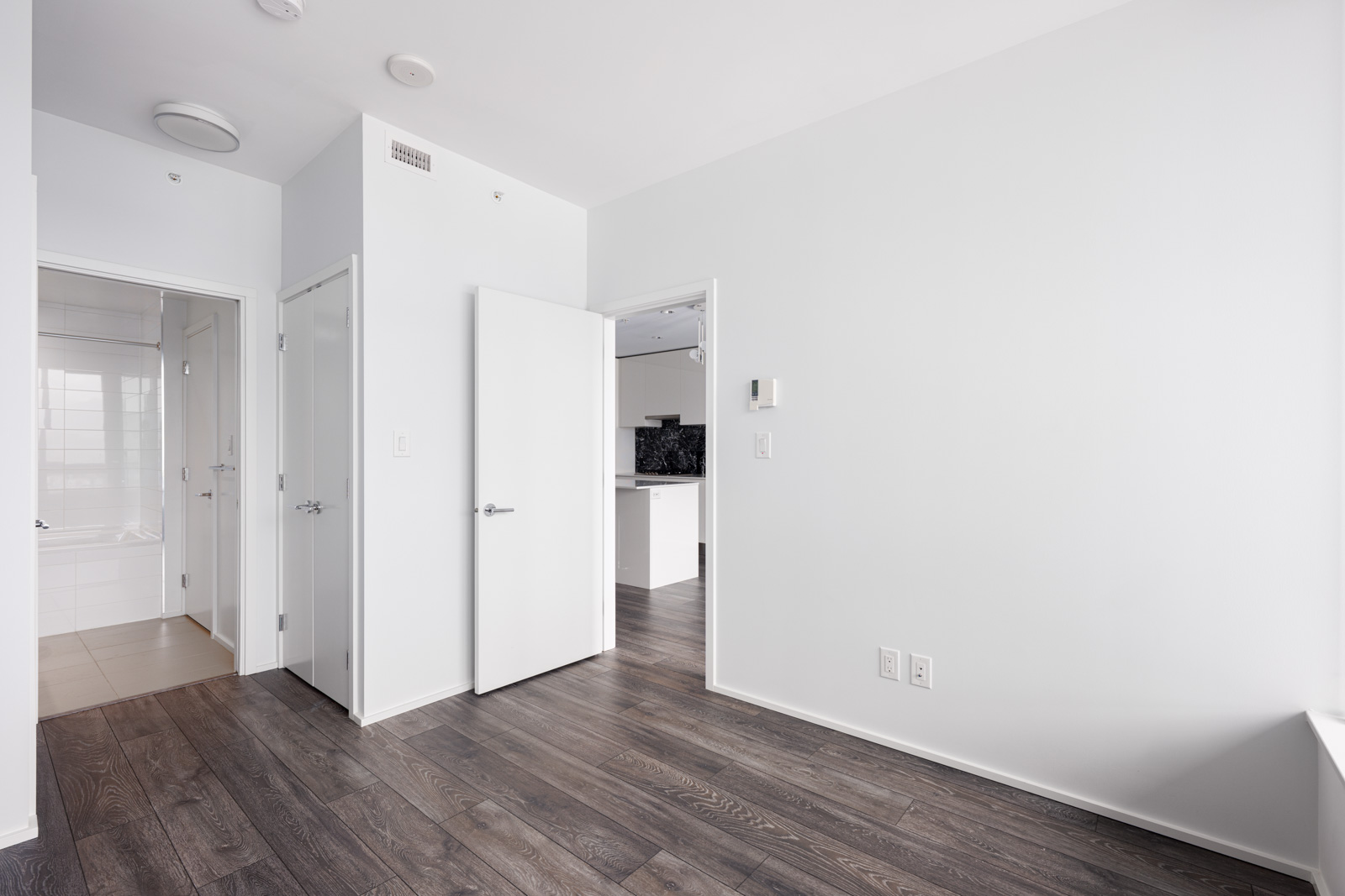 Empty, modern room with white walls, dark wood floor, open doors leading to a bathroom and kitchen, and ceiling lights visible.