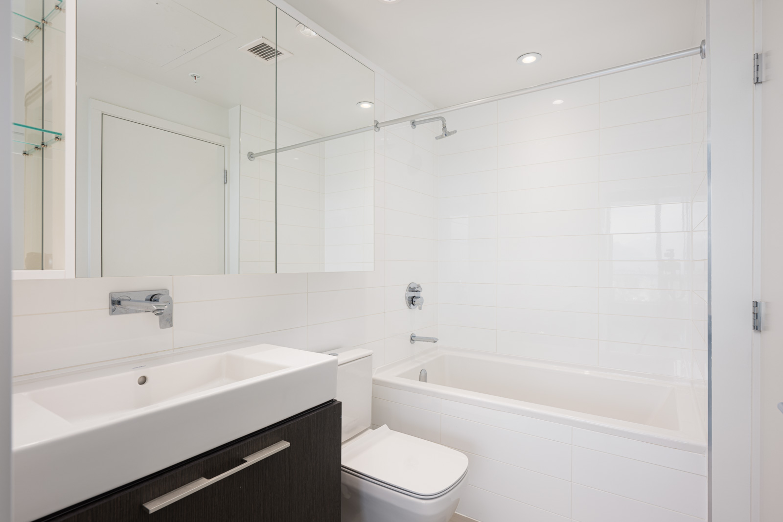 Modern white bathroom with a rectangular sink, dark vanity, toilet, and bathtub with a glass shower screen; recessed lighting and large mirror above the sink.