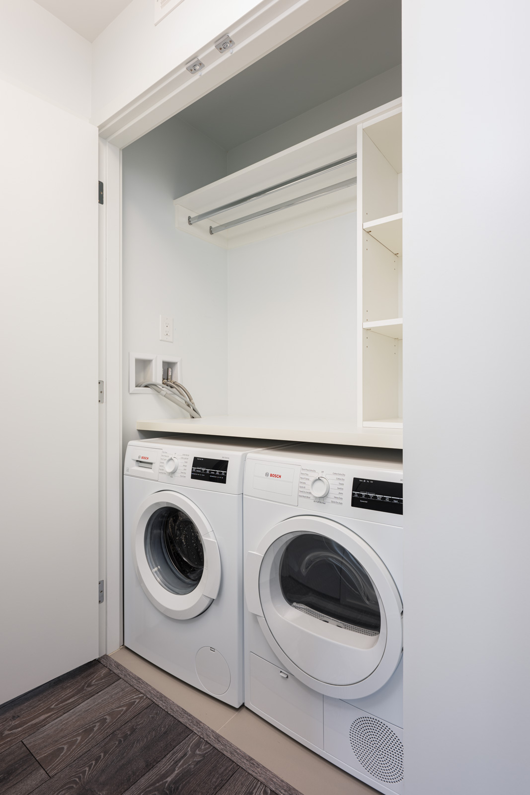 A compact laundry closet with a front-loading washing machine and dryer, open shelving, a hanging rail, and a white interior.