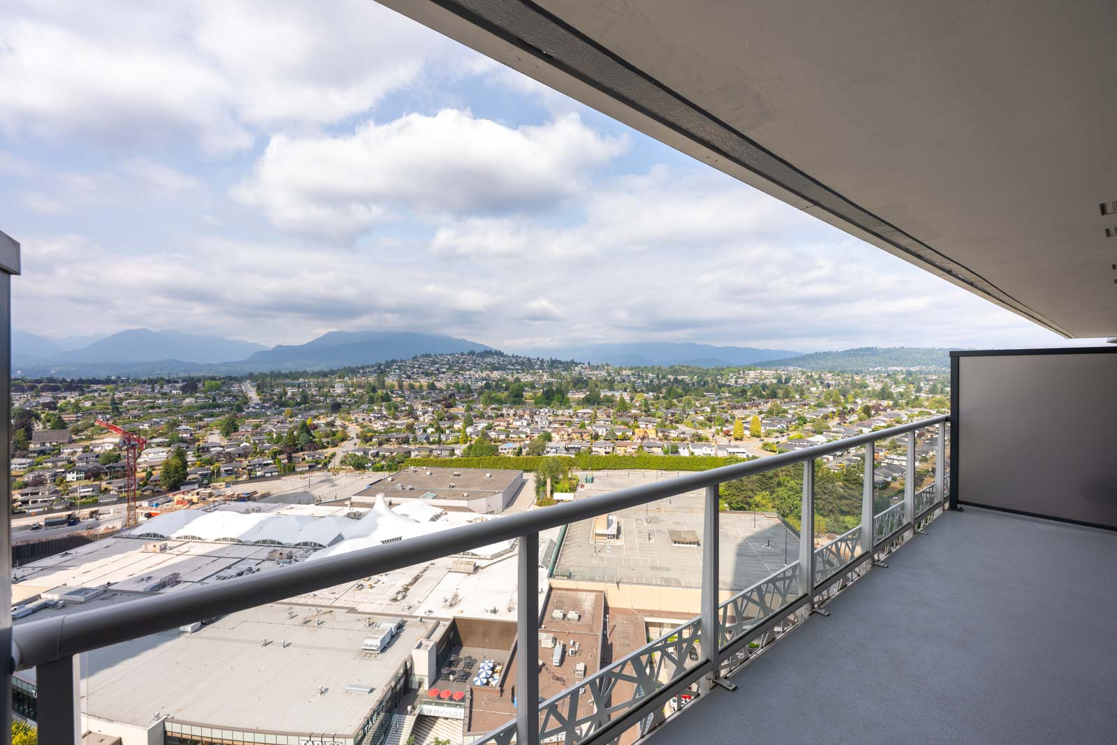 View from a high balcony overlooking a cityscape with buildings, trees, and distant mountains under a partly cloudy sky.