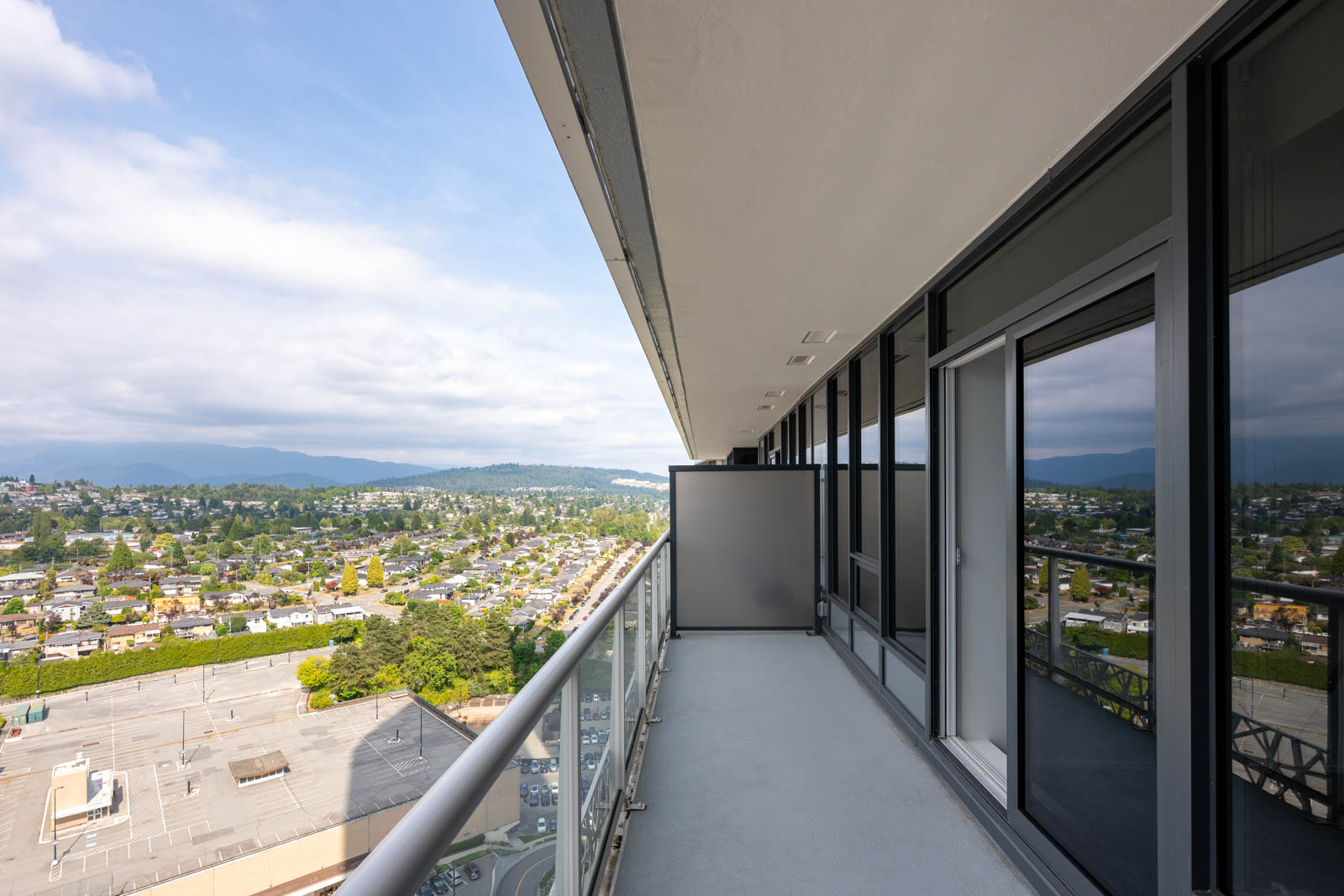 A high-rise balcony with glass railing overlooks a suburban area, visible under a partly cloudy sky. Reflective windows line the building on the right.