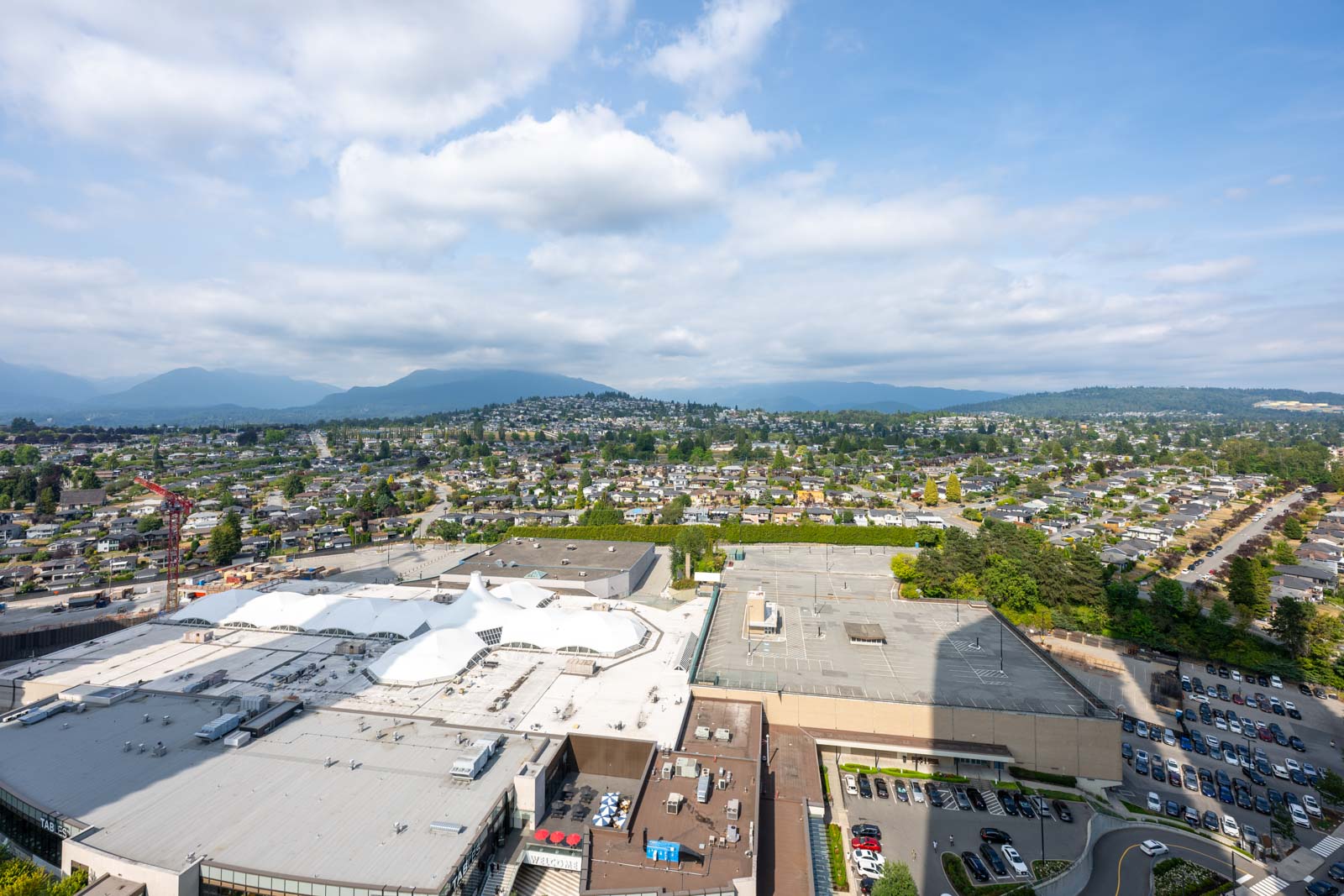 Aerial view of a commercial building with white rooftops in the foreground and a residential neighborhood, hills, and a partly cloudy sky in the background.