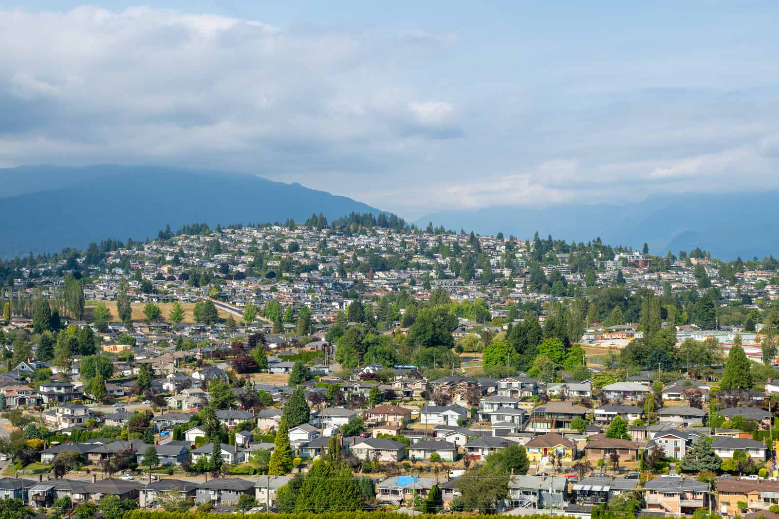 A suburban neighborhood with numerous houses and trees, set on and around a hill under a partly cloudy sky, with mountains in the distant background.